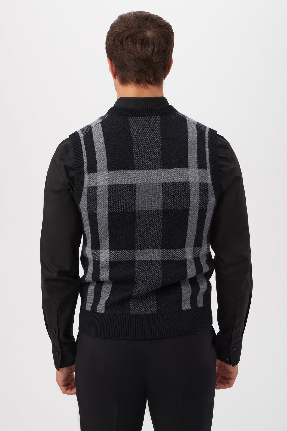 A man with short brown hair is seen from behind, wearing a black long-sleeve shirt and the Neso Plaid Knit Sweater Vest - Merino Wool in black and gray, standing against a plain white background.