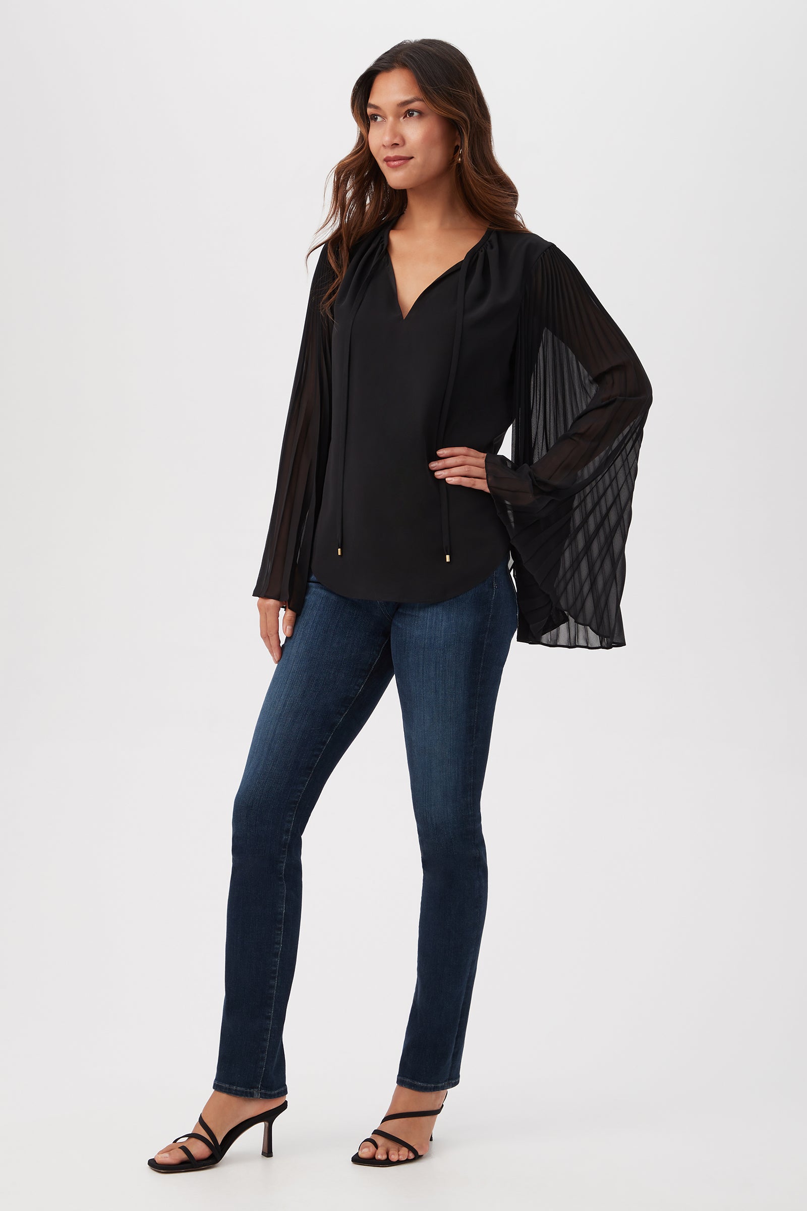 A woman poses against a plain background wearing the Golden Root Split V-Neck Top with accordion-pleated long sleeves, styled with blue jeans and black heeled sandals.