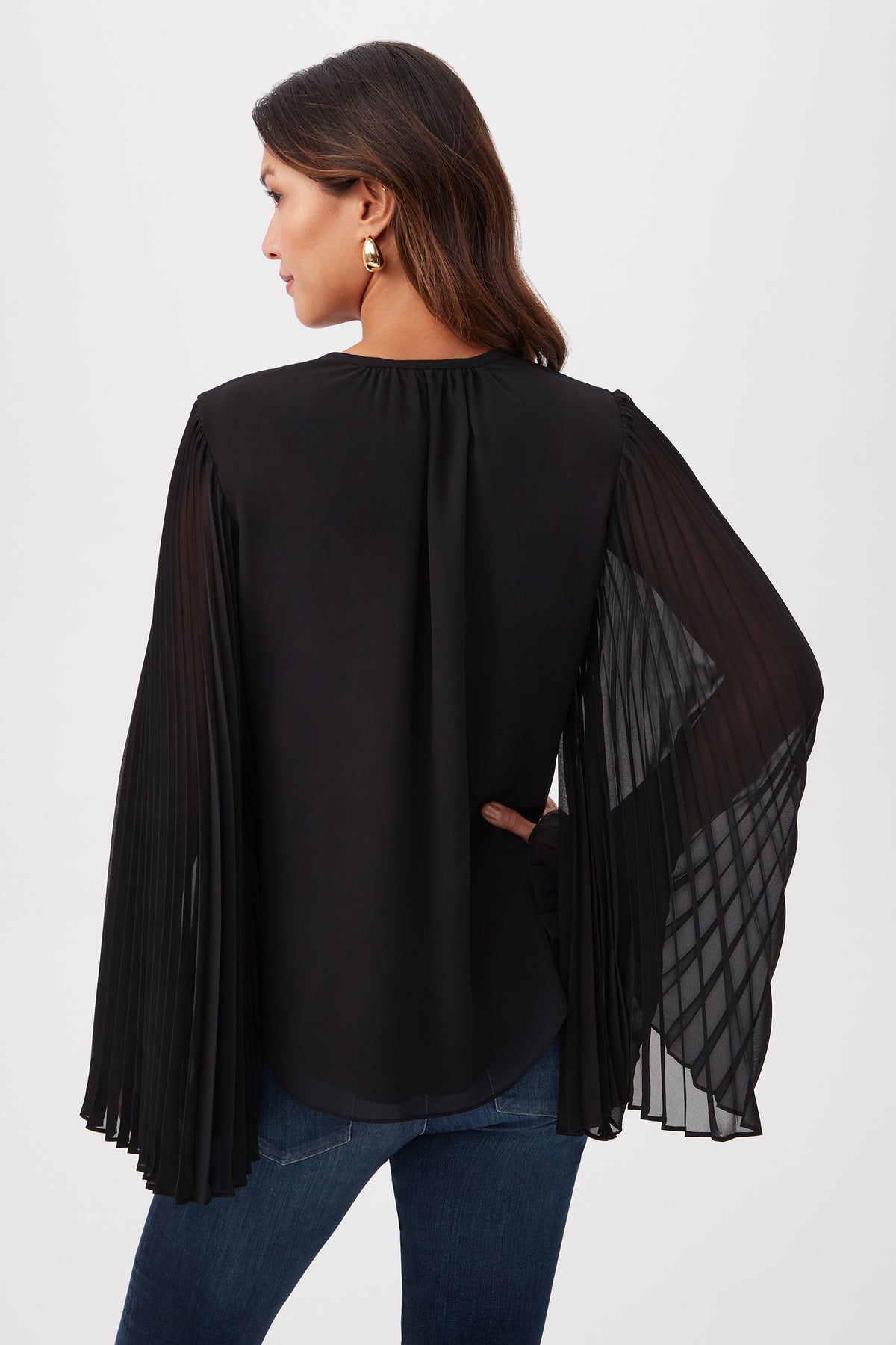 A woman with brown hair stands facing away, wearing the Golden Root Split V-Neck Top with accordion-pleated sheer long sleeves, blue jeans, and gold statement earrings. One hand rests on her hip.