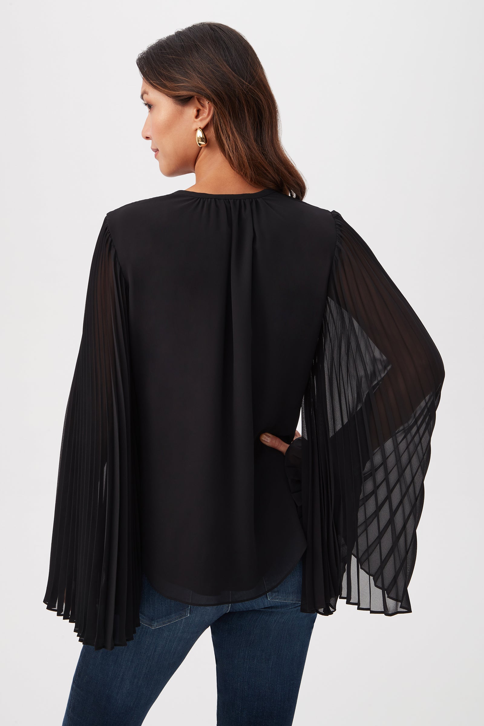 A woman with brown hair stands facing away, wearing the Golden Root Split V-Neck Top with accordion-pleated sheer long sleeves, blue jeans, and gold statement earrings. One hand rests on her hip.