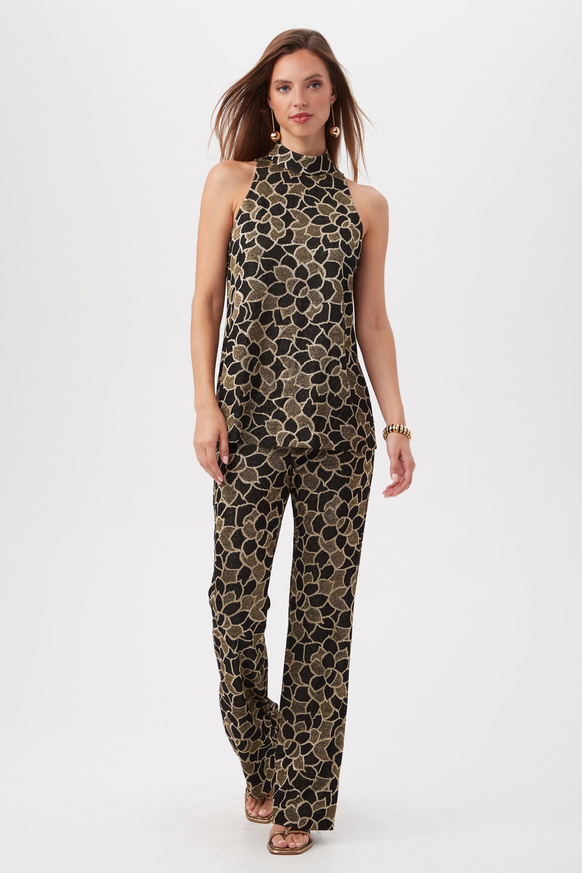 A woman walks confidently against a plain white background wearing Jacoba Metallic Knit Jacquard Bootcut Pants, gold earrings, a gold bracelet, and strappy heels.