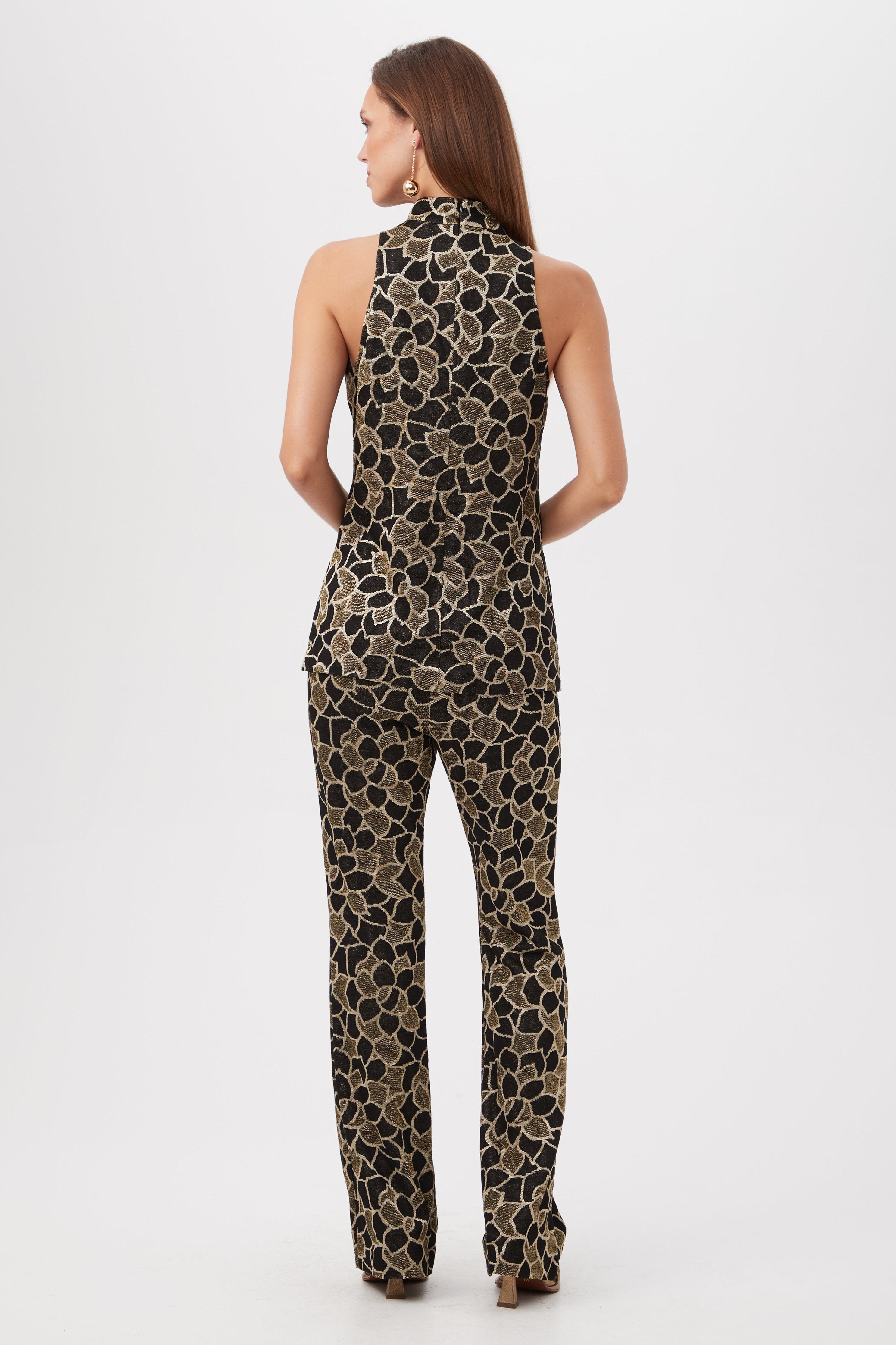 A woman faces away, wearing the Northern Lights Knit Jacquard Sleeveless Mockneck Tunic Top with a gold floral pattern and wide-leg black pants, set against a plain white background.