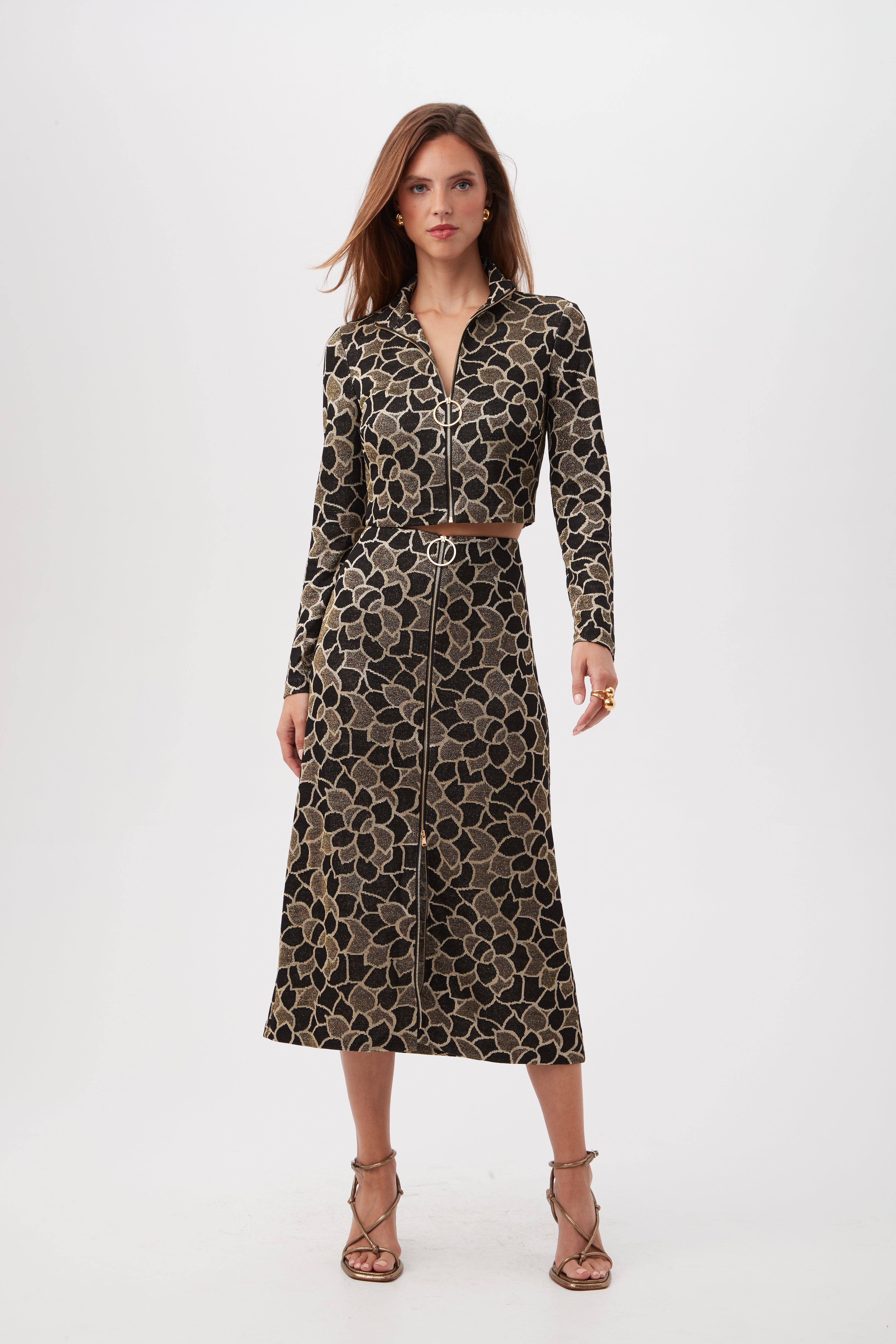 A woman stands against a white background wearing the Frosty Meadows Cropped Knit Jacquard Zip-Front Top with long sleeves and a floral pattern, her hair loose and paired with strappy high-heeled sandals.