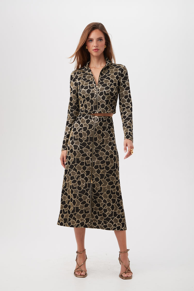 A woman stands against a white background wearing the Frosty Meadows Cropped Knit Jacquard Zip-Front Top with long sleeves and a floral pattern, her hair loose and paired with strappy high-heeled sandals.