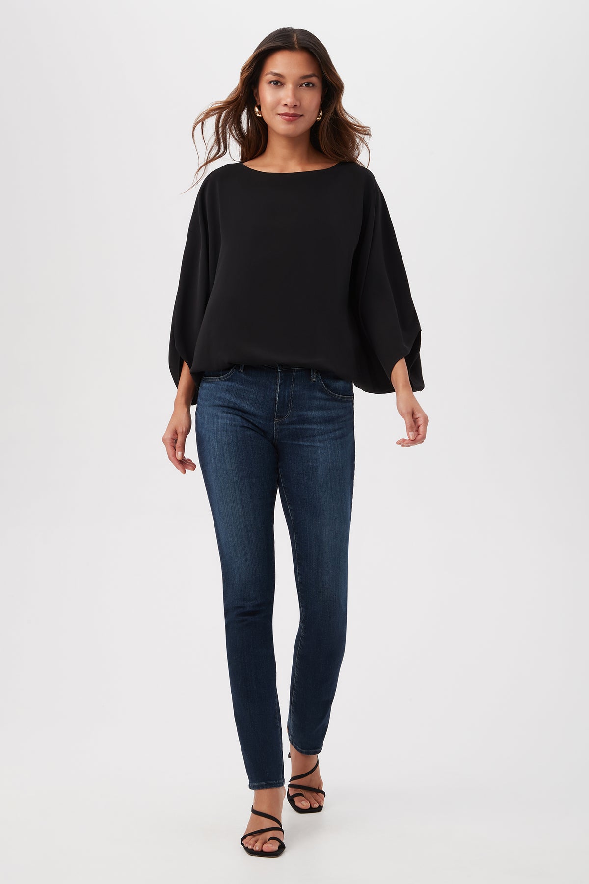 A woman walks confidently in the Manhattan Relaxed Short Dolman Sleeve Top, dark blue skinny jeans, and black sandals against a plain white background.