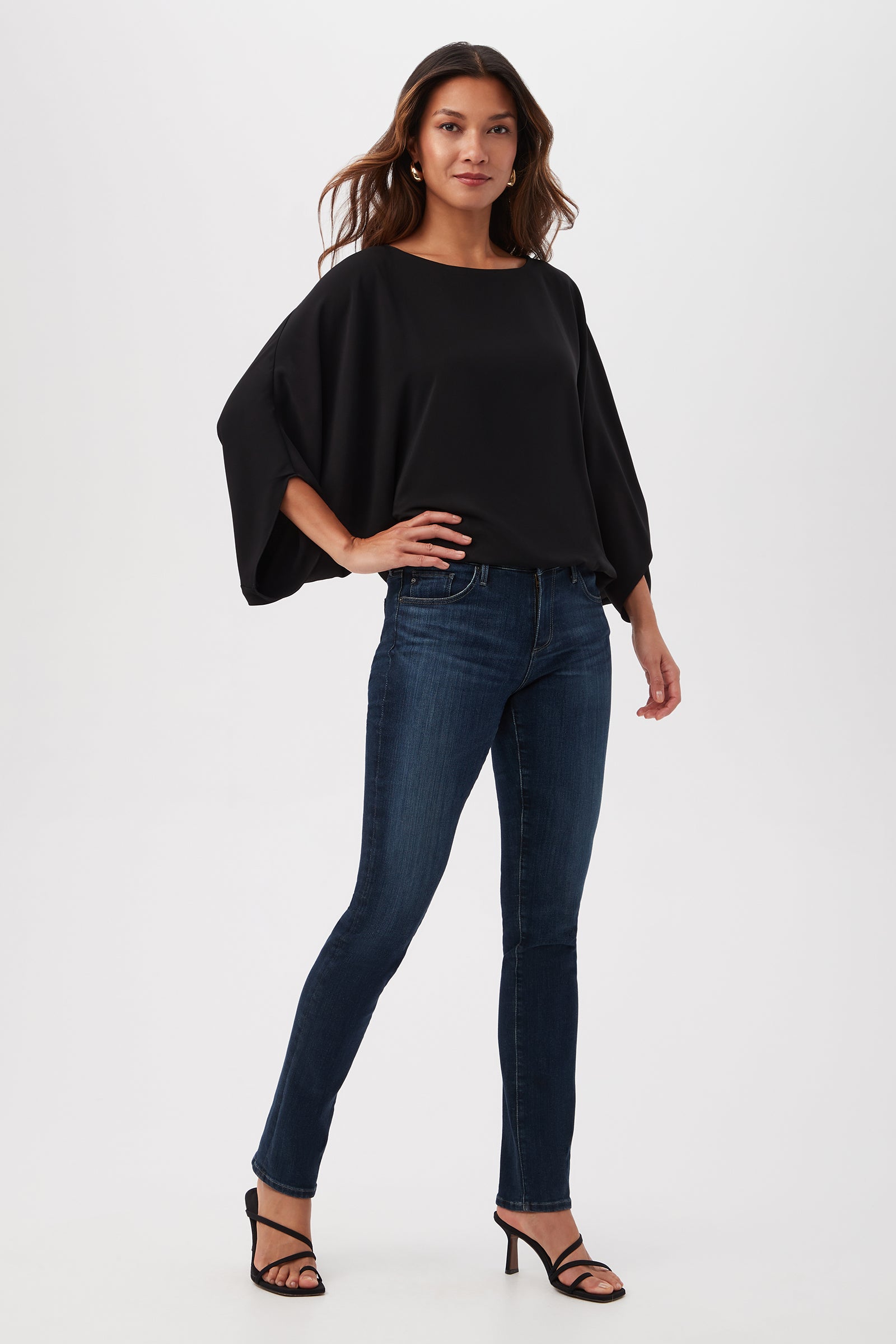 A woman stands confidently against a plain background, wearing the Manhattan Relaxed Short Dolman Sleeve Top with dark blue skinny jeans and black high-heeled sandals. She has long, wavy hair and a relaxed expression.