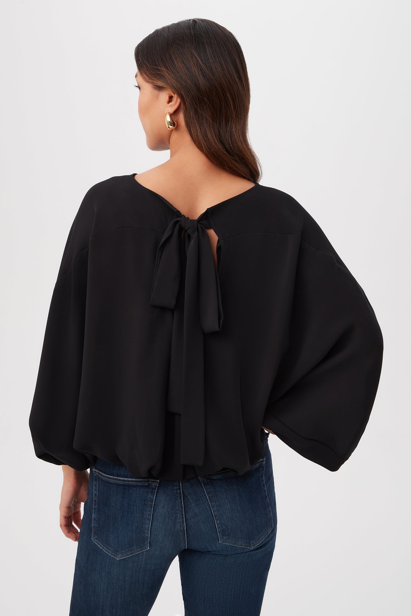 A woman with long dark hair, gold hoop earrings, and blue jeans stands facing away from the camera in a black Manhattan Relaxed Short Dolman Sleeve Top with a large back bow against a plain white background.