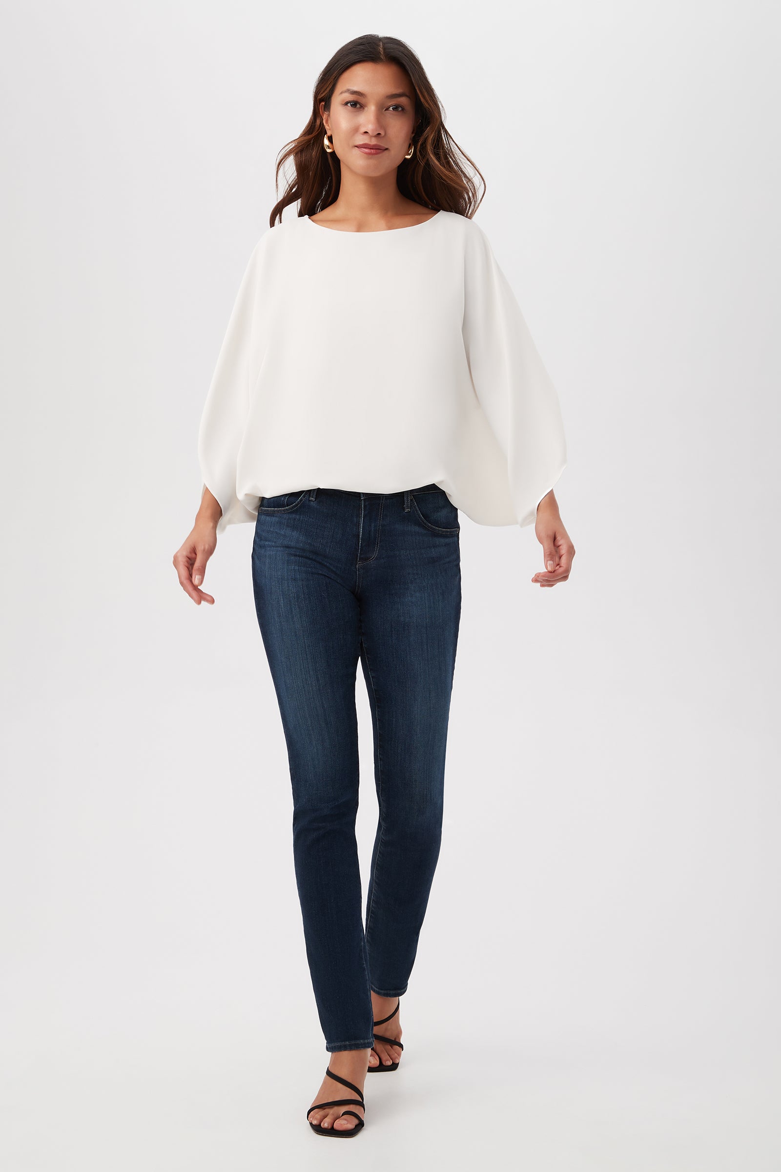 A woman wears the Manhattan Relaxed Short Dolman Sleeve Top, styled with dark blue skinny jeans and black sandals, as she walks against a plain white background.