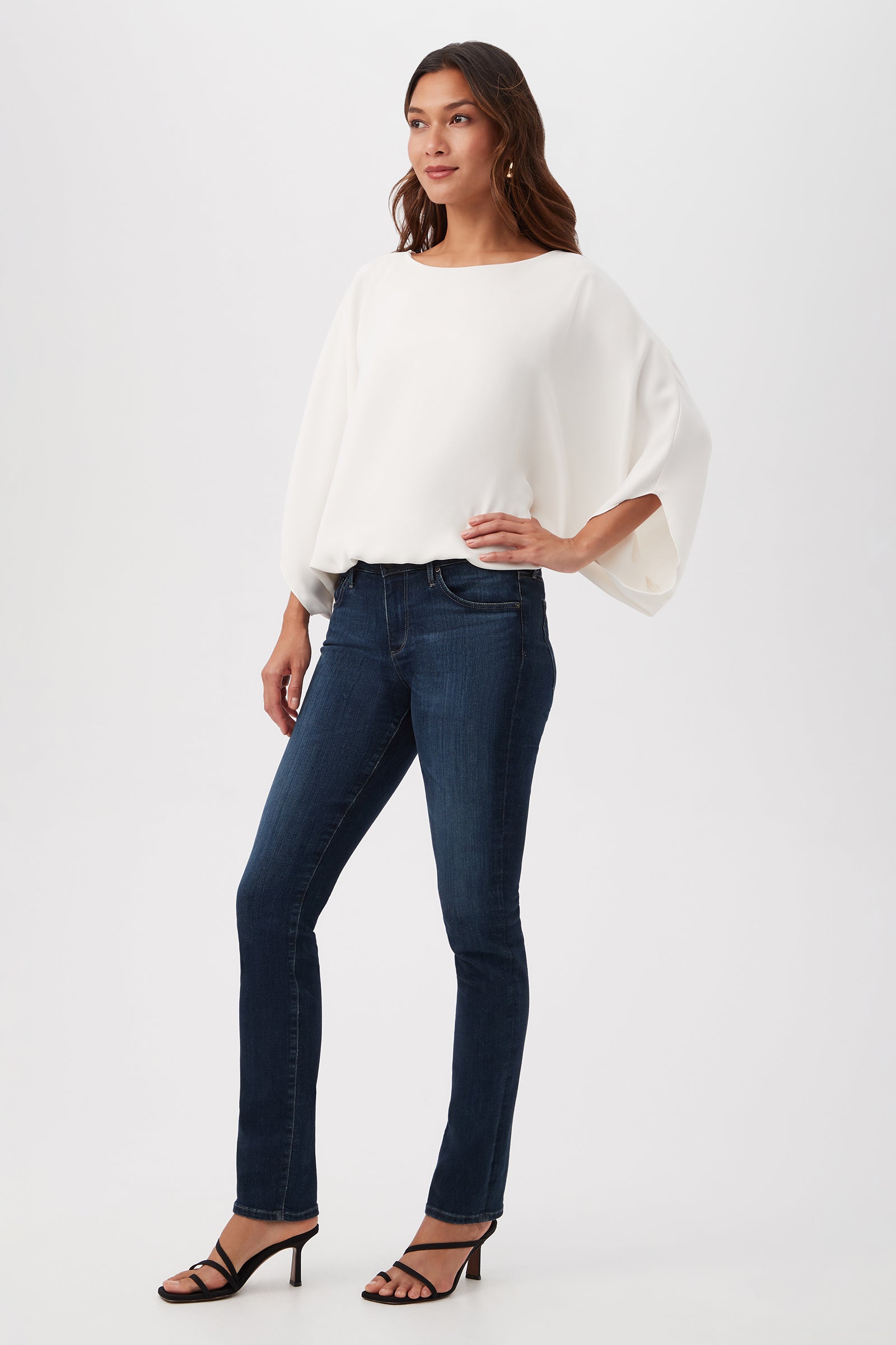 A woman with long wavy brown hair stands against a plain backdrop wearing the Manhattan Relaxed Short Dolman Sleeve Top, dark blue skinny jeans, and black strappy high-heeled sandals, with one hand resting on her hip.