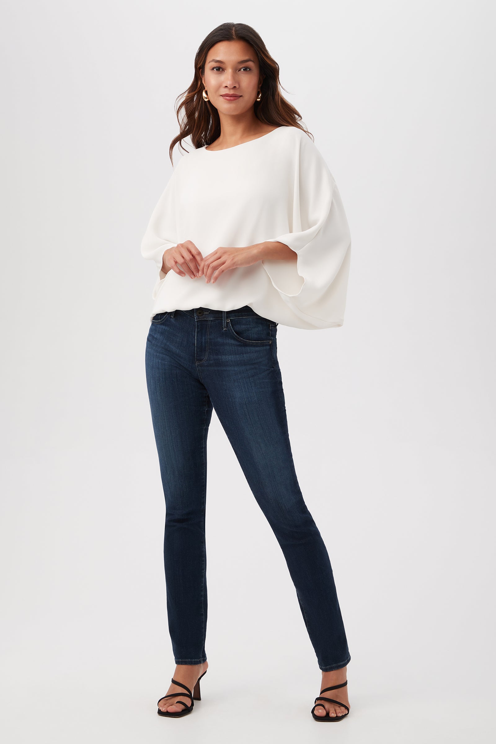 A woman poses confidently against a white background in the Manhattan Relaxed Short Dolman Sleeve Top, paired with dark blue skinny jeans and black strappy heels. Her long wavy hair complements her relaxed and stylish look.