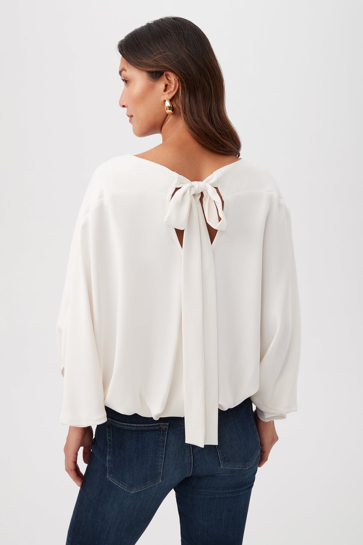 A woman with long brown hair, seen from behind against a plain light background, wears the Manhattan Relaxed Short Dolman Sleeve Top— a loose white blouse featuring a large bow at the back neckline—paired with dark blue jeans.
