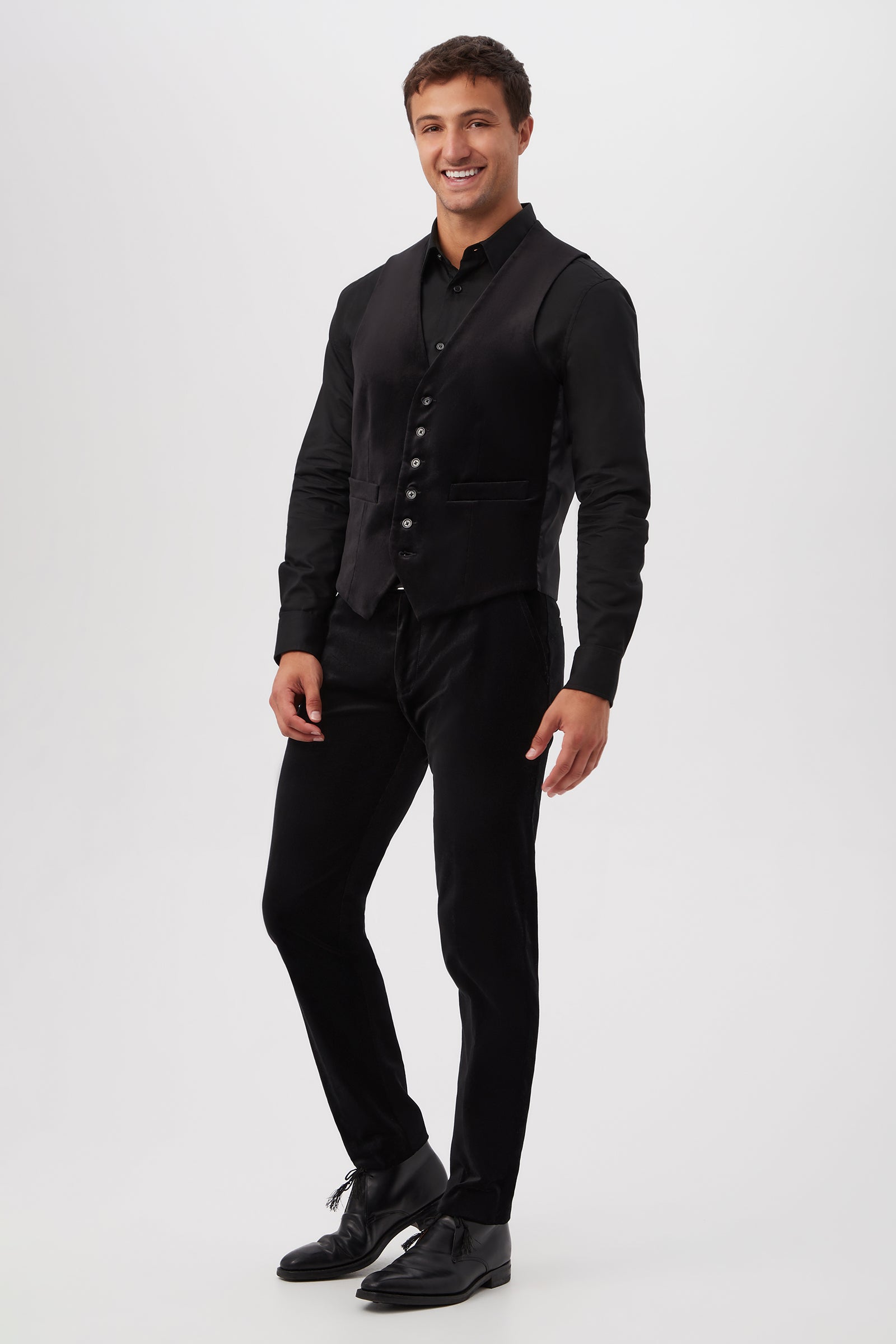 A man smiles against a plain white background, wearing the Irving Vest in Black Velveteen over a black shirt, paired with black pants and black shoes.