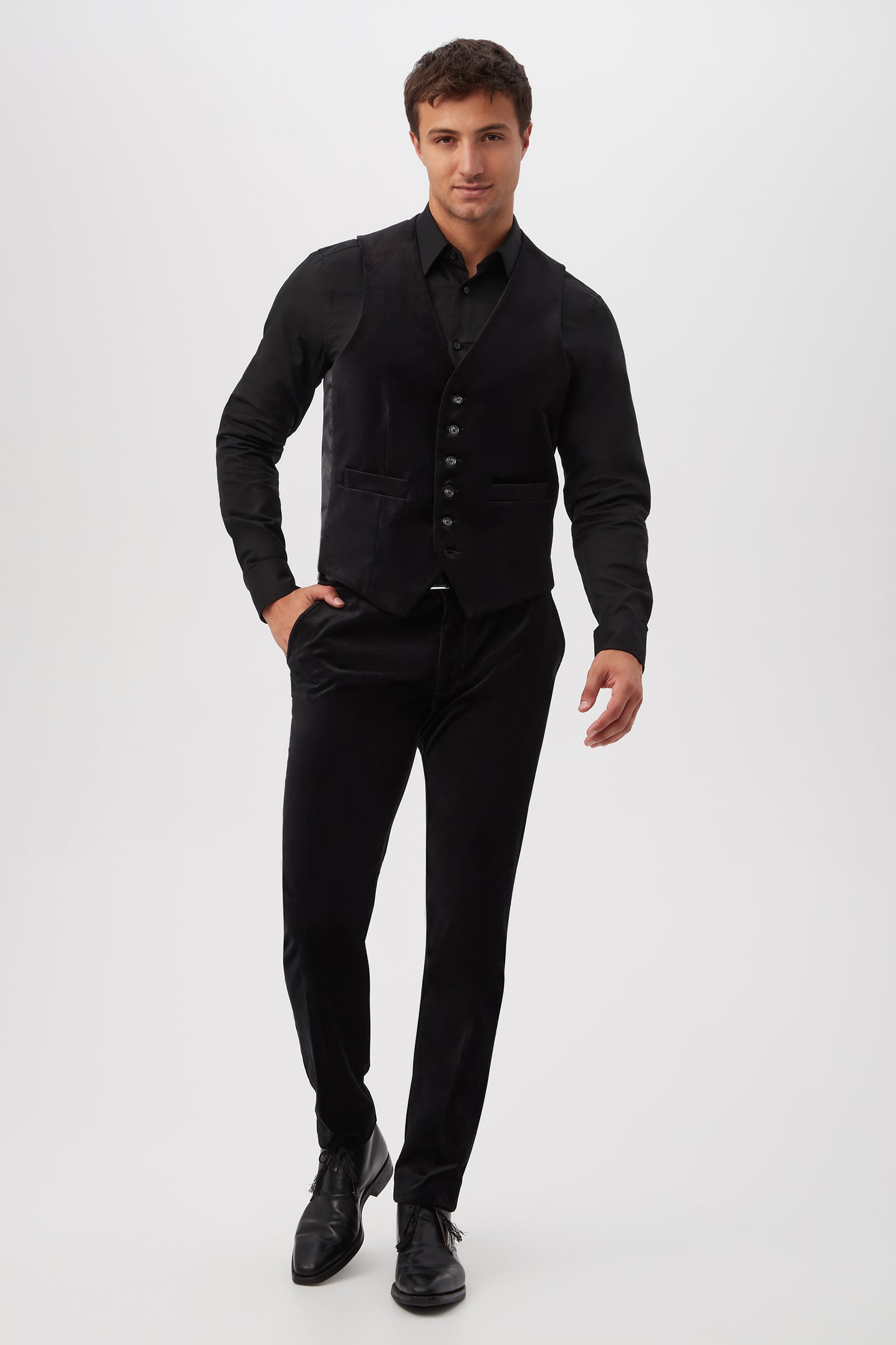 A man wearing the Irving Vest - Black Velveteen stands against a plain white background, dressed in all black with one hand in his pocket, smiling slightly as he walks forward.