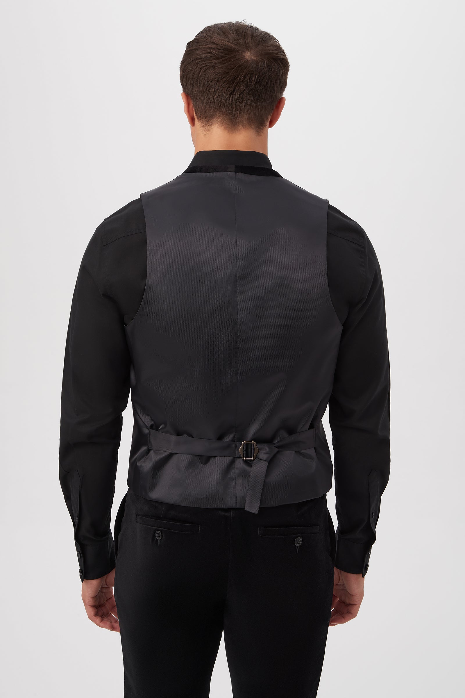 A man is seen from behind wearing the Irving Vest in black velveteen, paired with a black dress shirt and trousers; the vest features an adjustable back strap and buckle.
