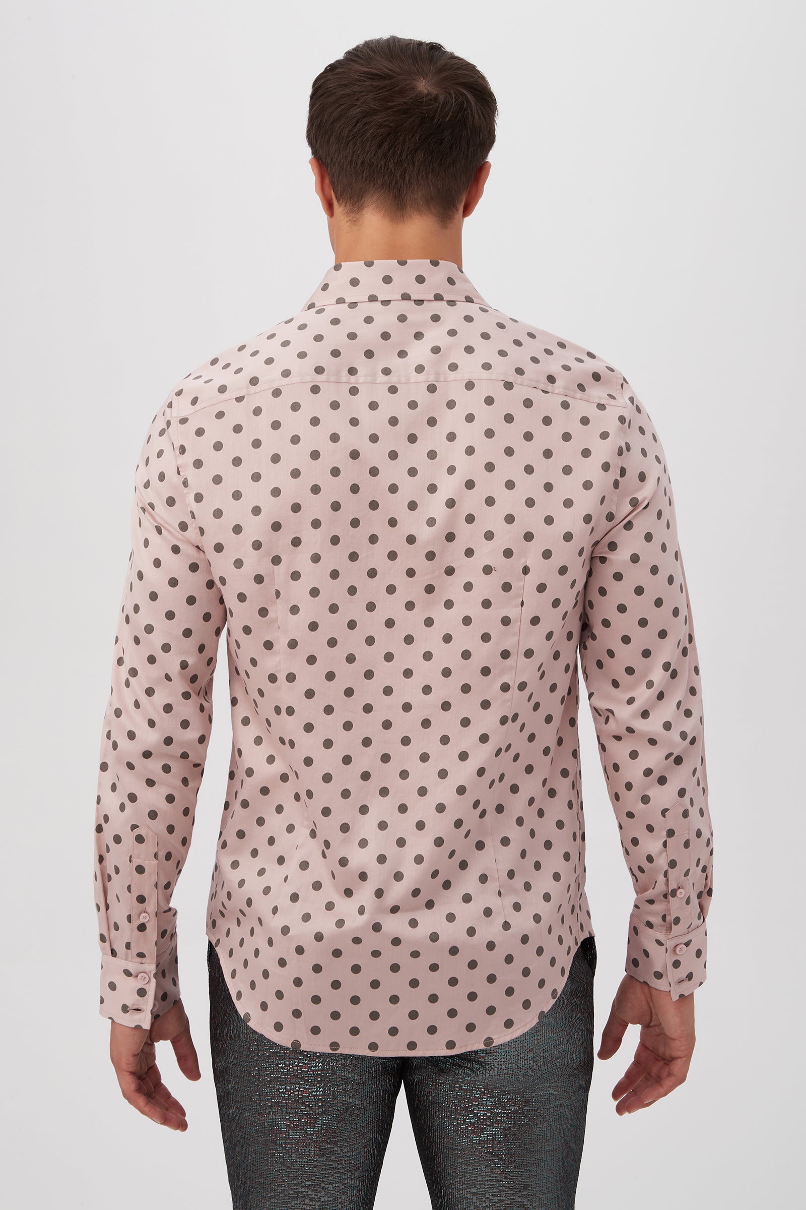 A man stands facing away, wearing the Aaron Slim Fit Long-Sleeve Shirt in light pink with black polka dots and shiny dark pants. The background is plain white.