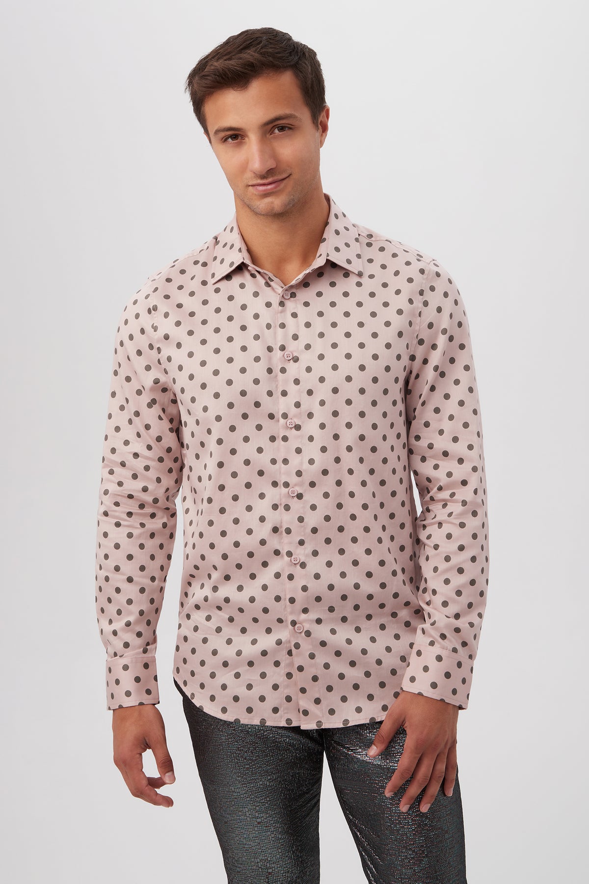 A man in the Aaron Slim Fit Long-Sleeve Shirt, light pink with black polka dots, and shiny dark textured pants stands against a plain white background, smiling softly.