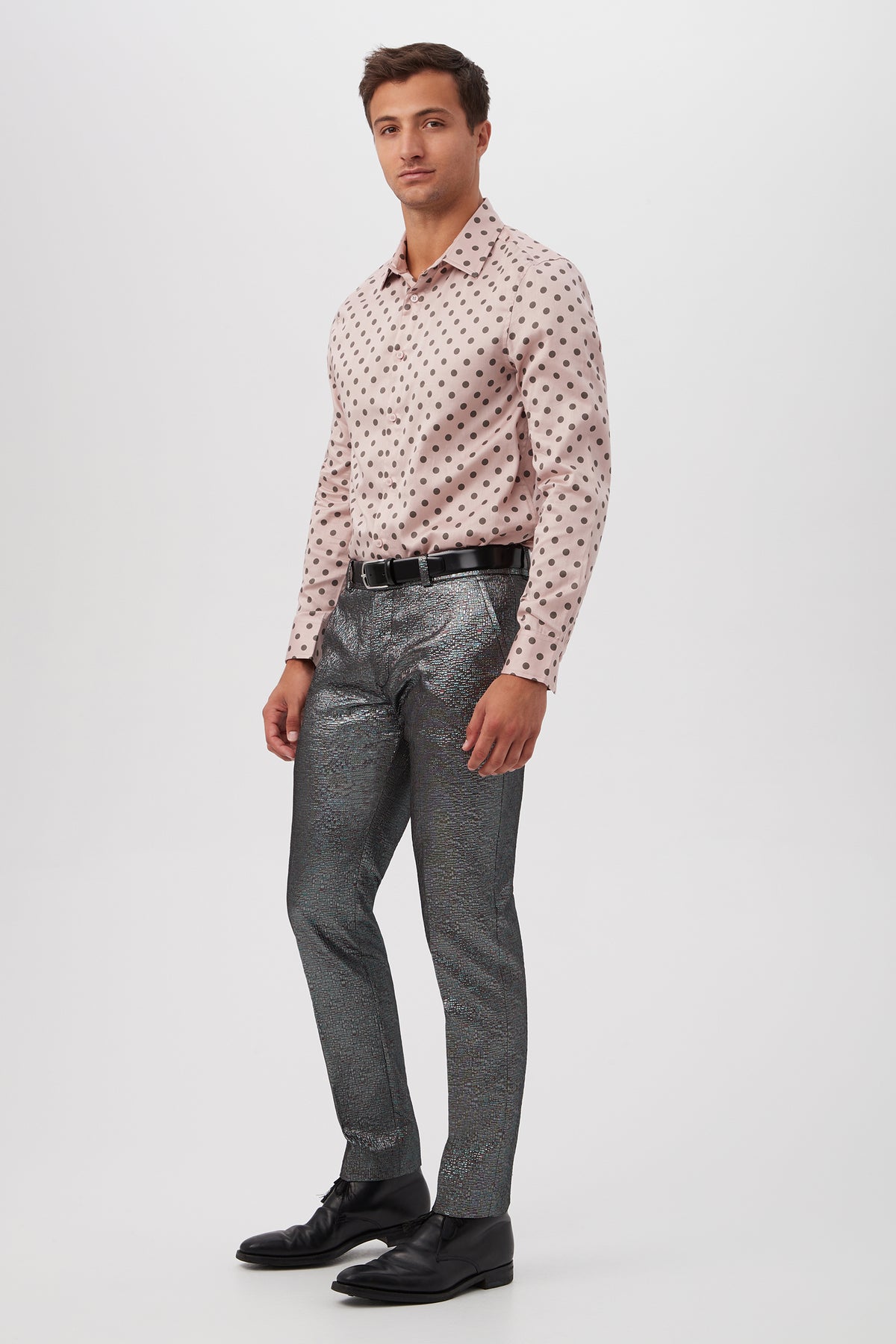 A man stands against a plain white background wearing the Aaron Slim Fit Long-Sleeve Shirt in light pink with dark polka dots, metallic silver textured pants, a black belt, and black dress shoes.