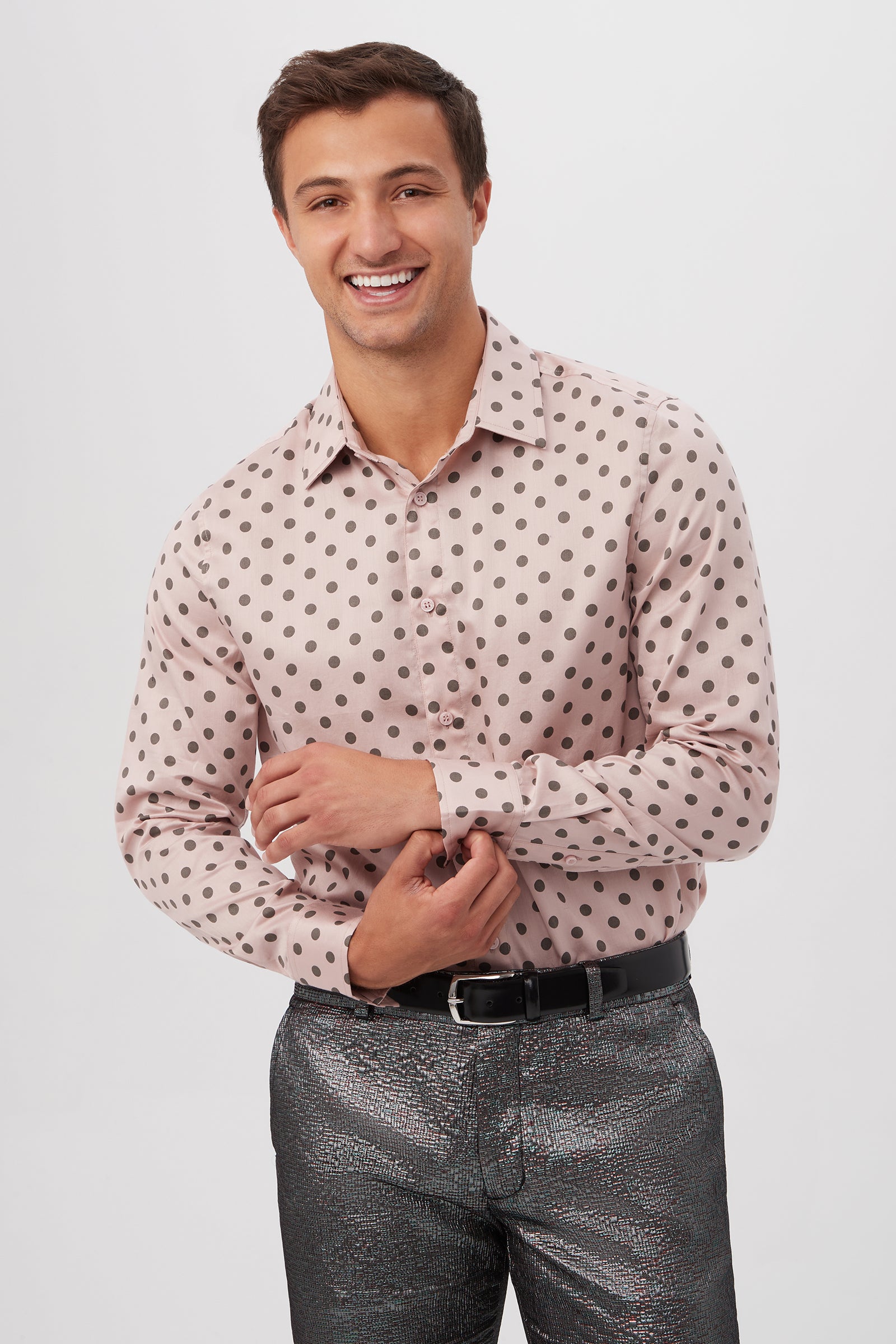 A smiling man stands against a plain background, wearing the Aaron Slim Fit Long-Sleeve Shirt in Dusty Rose with black polka dots and shiny metallic trousers with a black belt.