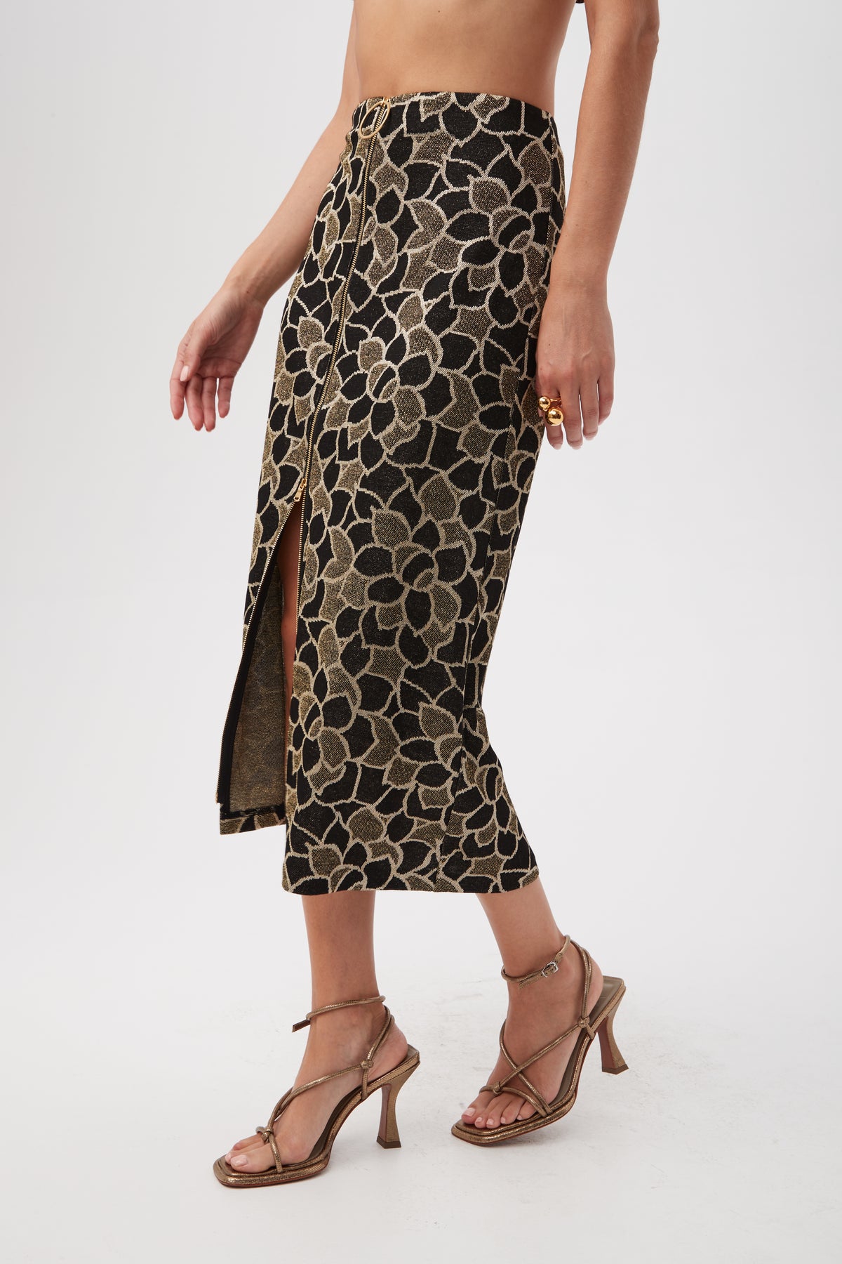 A person models the Zane Metallic Knit Jacquard Zip-Front Midi Skirt, featuring a gold and black floral pattern and side slit, paired with heeled sandals. The upper body is partially cropped against a plain white background.