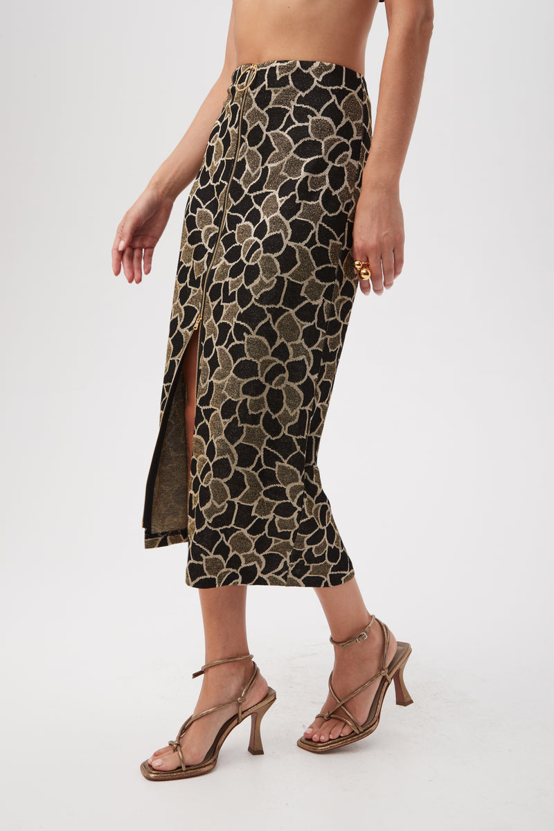A person models the Zane Metallic Knit Jacquard Zip-Front Midi Skirt, featuring a gold and black floral pattern and side slit, paired with heeled sandals. The upper body is partially cropped against a plain white background.