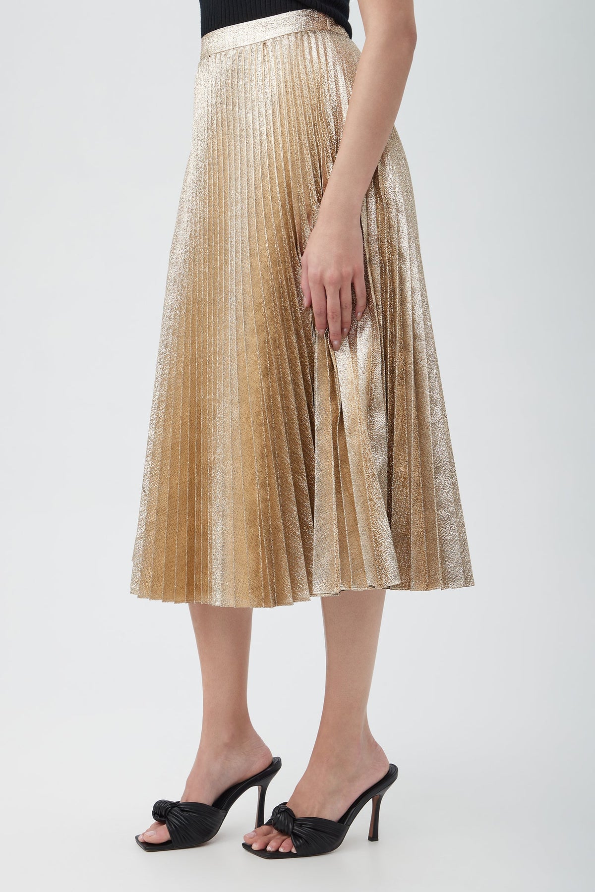 A woman stands against a plain light background wearing the Faye Luminary Lamé Midi Skirt With Accordion Pleats, paired with a black sleeveless top and black high-heeled sandals.