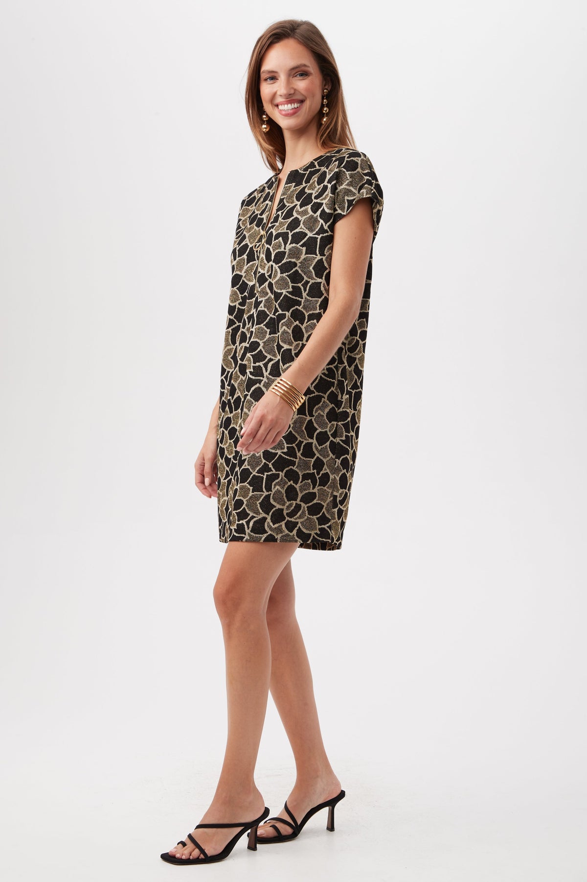 A woman stands sideways, smiling, wearing the Hasil 2 Short-Sleeve Shift Dress—a short black dress with a gold floral pattern—paired with black heeled sandals, hoop earrings, and bracelets against a plain white background.