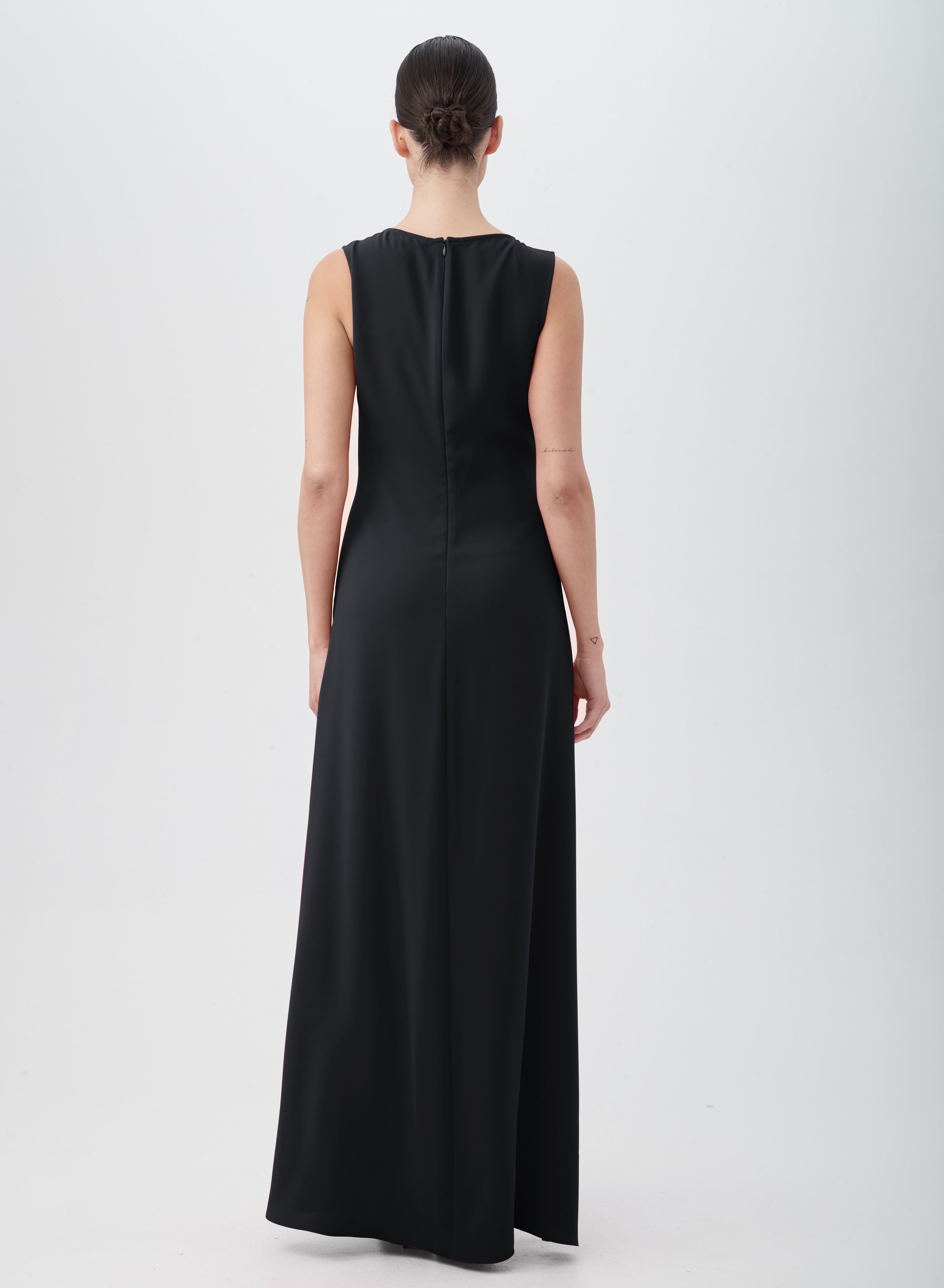 A person with a low bun stands facing away, wearing the floor-length, sleeveless MYTHICAL DRESS in black, made from 100% polyester. The background is plain and light-colored.
