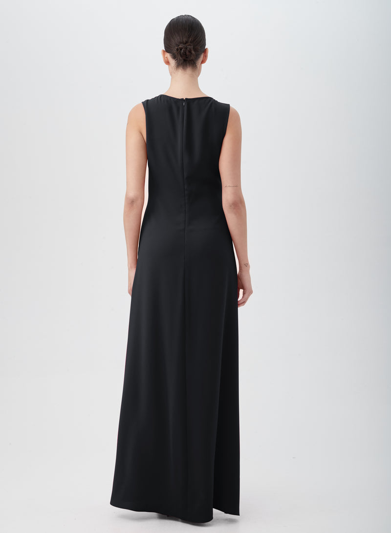 A person with a low bun stands facing away, wearing the floor-length, sleeveless MYTHICAL DRESS in black, made from 100% polyester. The background is plain and light-colored.