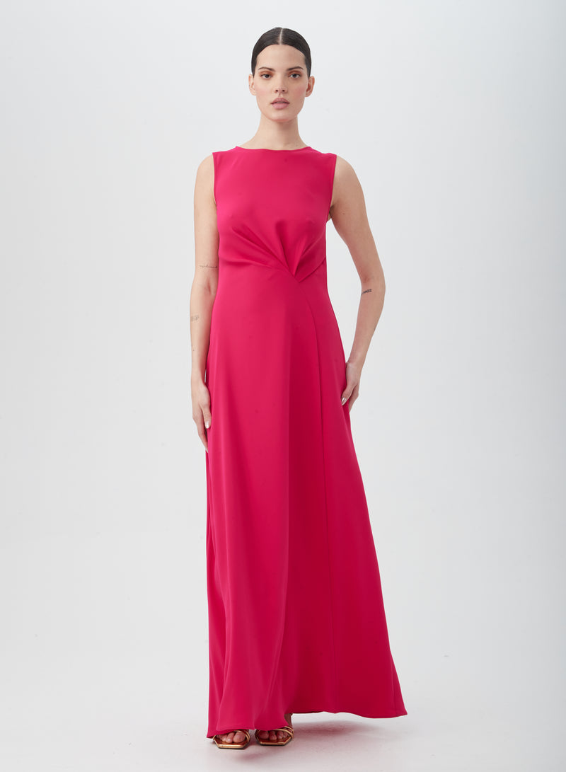 A woman stands against a plain background, wearing the MYTHICAL DRESS—a floor-length, sleeveless, bright pink gown crafted from silky polyester with subtle waist pleating. Her hair is pulled back as she looks directly at the camera.