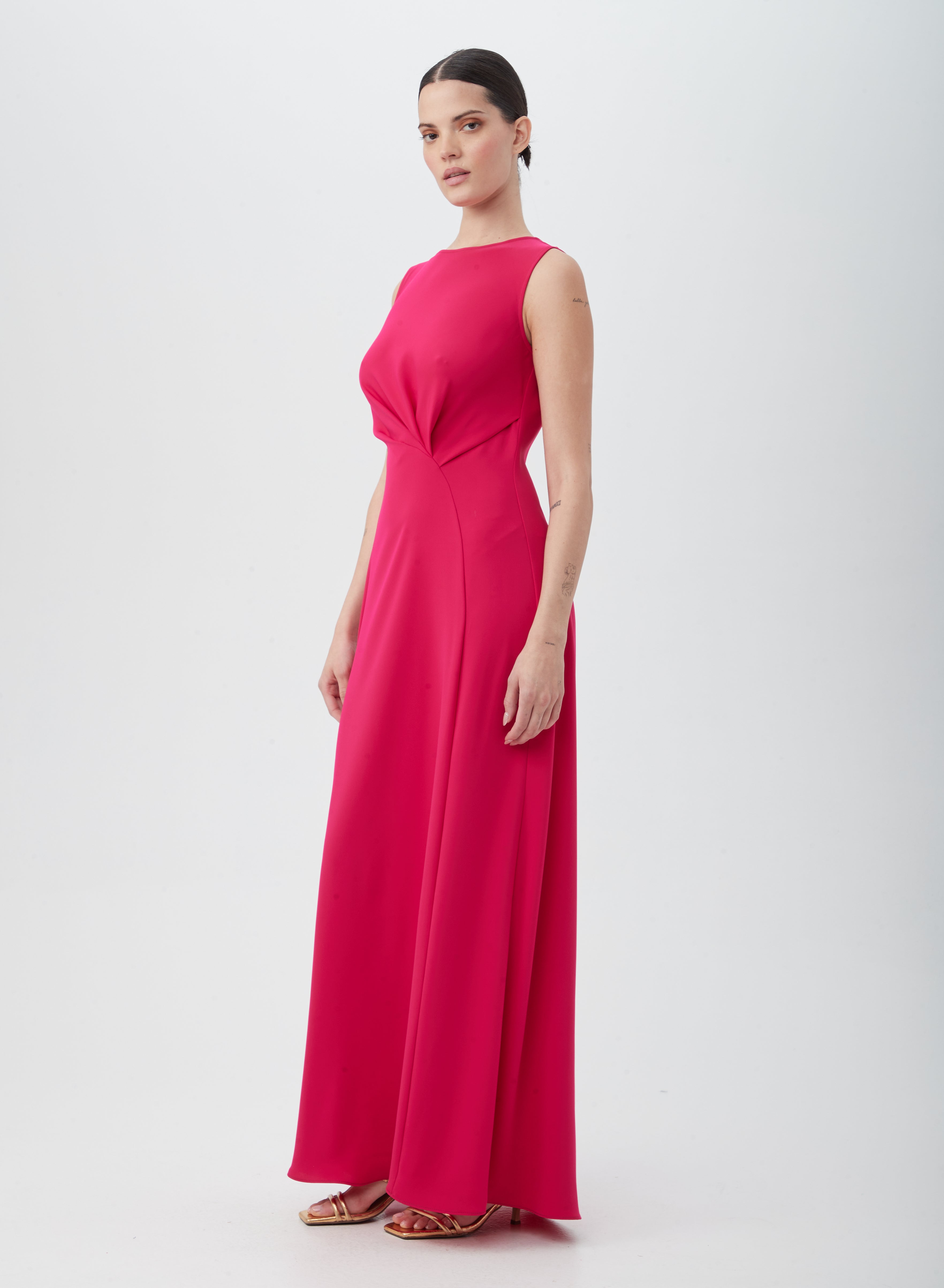 A woman poses against a white background in the MYTHICAL DRESS, a sleeveless, floor-length bright pink dress made of 100% polyester with a fitted bodice and flowing skirt, styled with gold strappy heels.