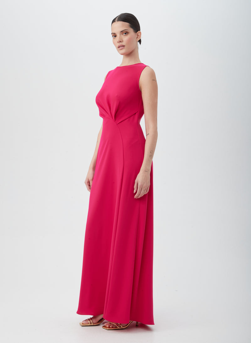 A woman poses against a white background in the MYTHICAL DRESS, a sleeveless, floor-length bright pink dress made of 100% polyester with a fitted bodice and flowing skirt, styled with gold strappy heels.