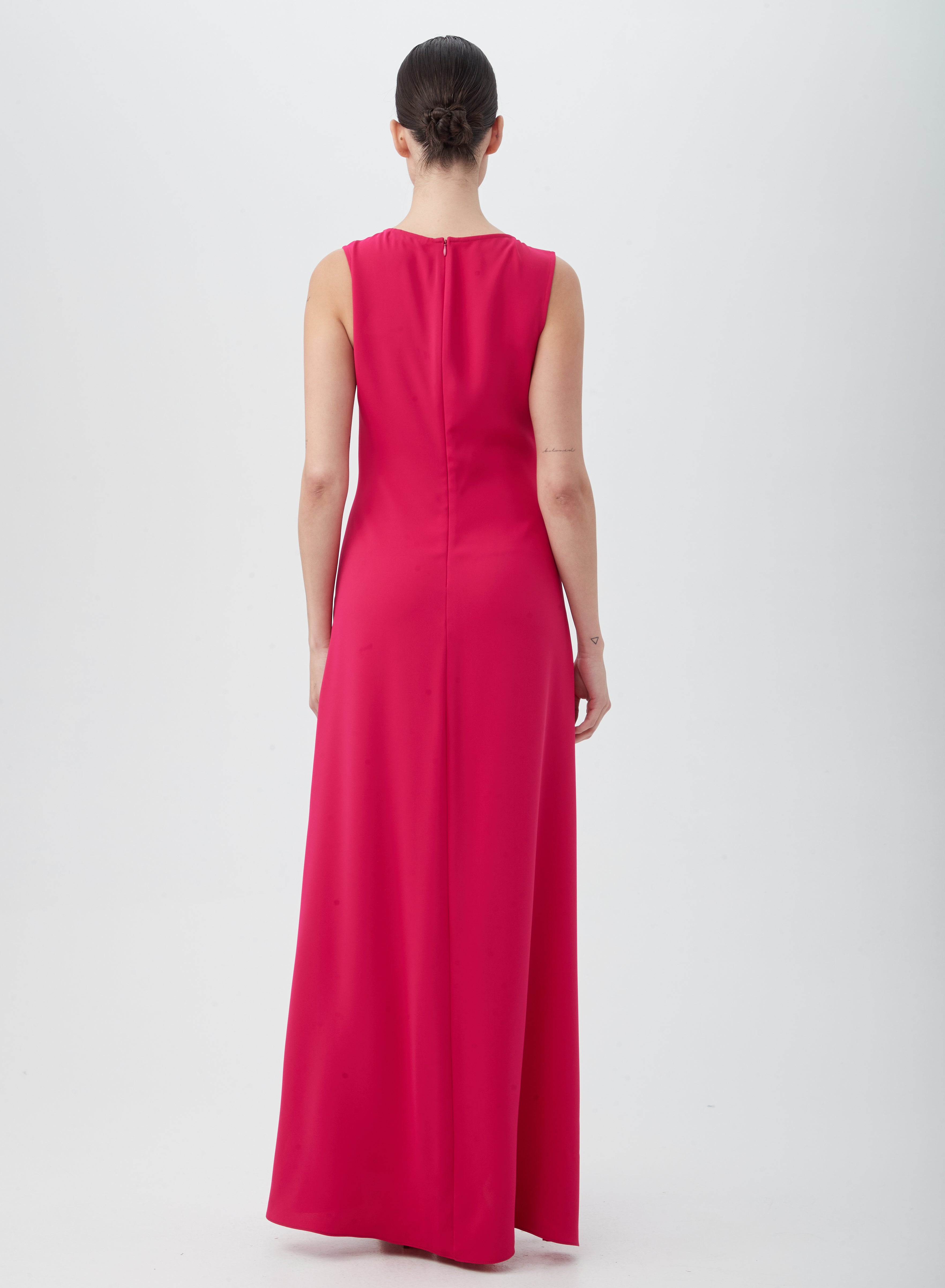 A woman with dark hair in a bun faces away, wearing the MYTHICAL DRESS—a sleeveless, floor-length bright pink dress made from 100% polyester. She stands against a plain white background.
