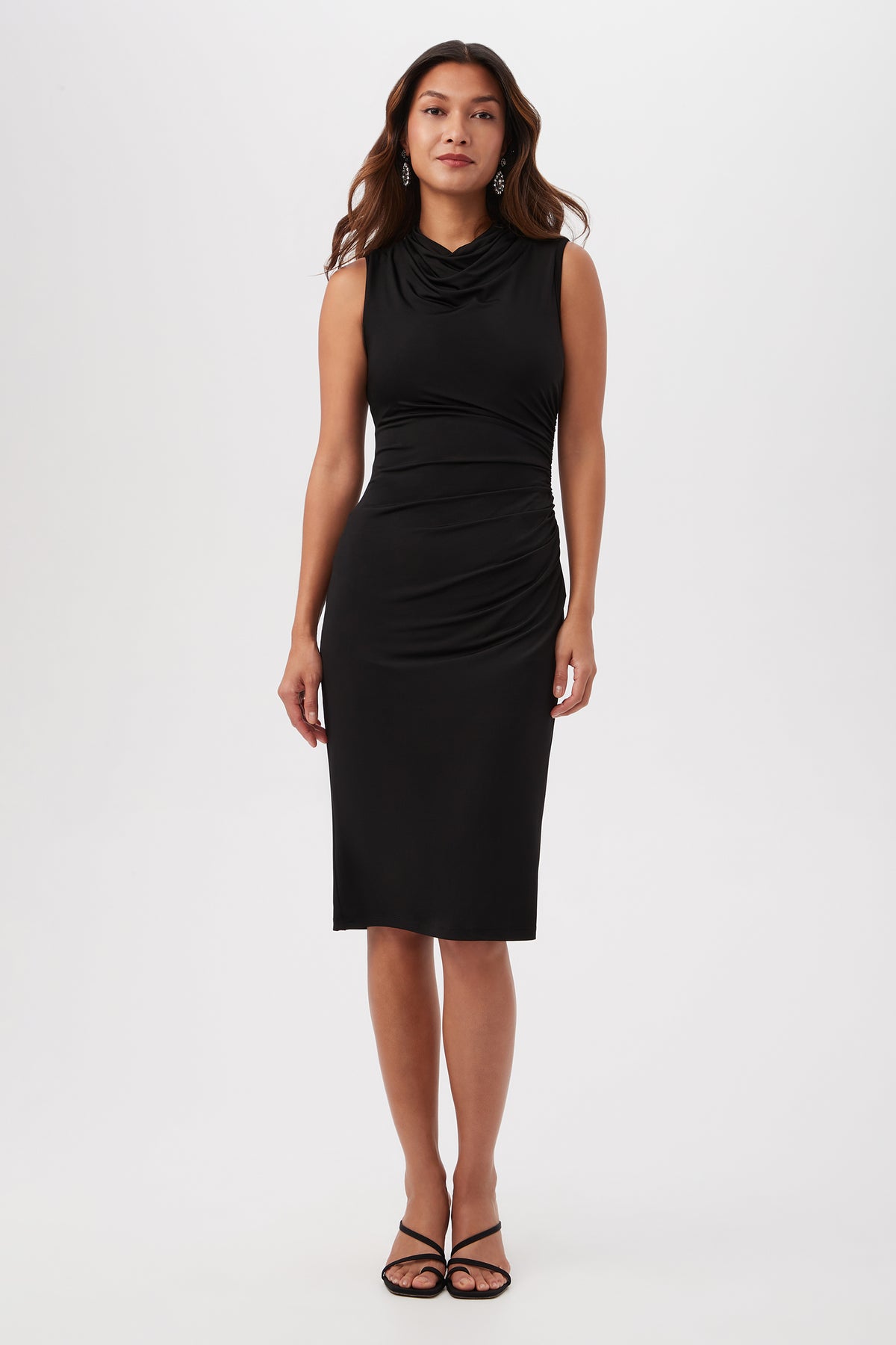 A woman faces forward in the black Spruce Sleeveless Slinky Jersey Midi Dress with a cowl neck and side ruching, paired with black strappy sandals, set against a plain white background.