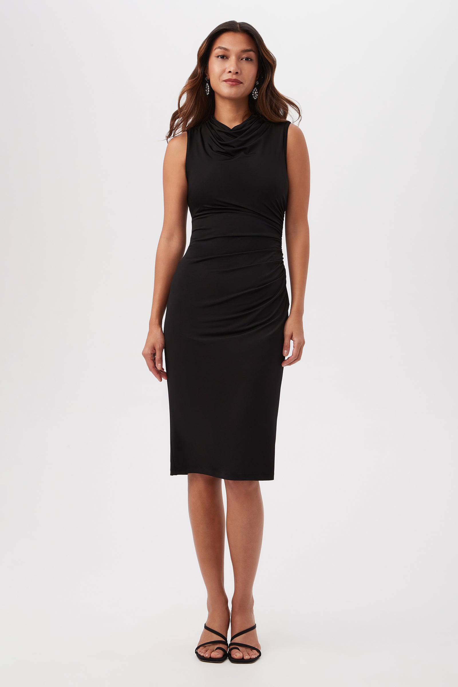 A woman faces forward in the black Spruce Sleeveless Slinky Jersey Midi Dress with a cowl neck and side ruching, paired with black strappy sandals, set against a plain white background.