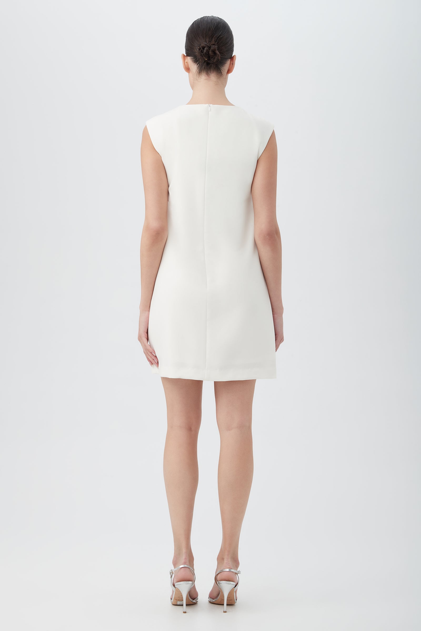 A woman faces away, wearing the Ruth Sleeveless Shift Dress With Decorative Front Detail in white and silver high-heeled sandals. Her hair is neatly pulled back as she poses against a plain light background.