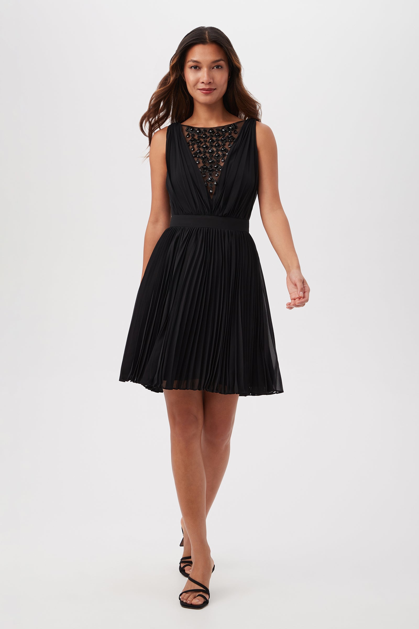 A woman with long brown hair smiles slightly as she walks forward in the Zathar Sleeveless Dress With Jeweled Front And Accordion Pleats, black strappy heels, against a plain white background. The dress is black and knee-length.