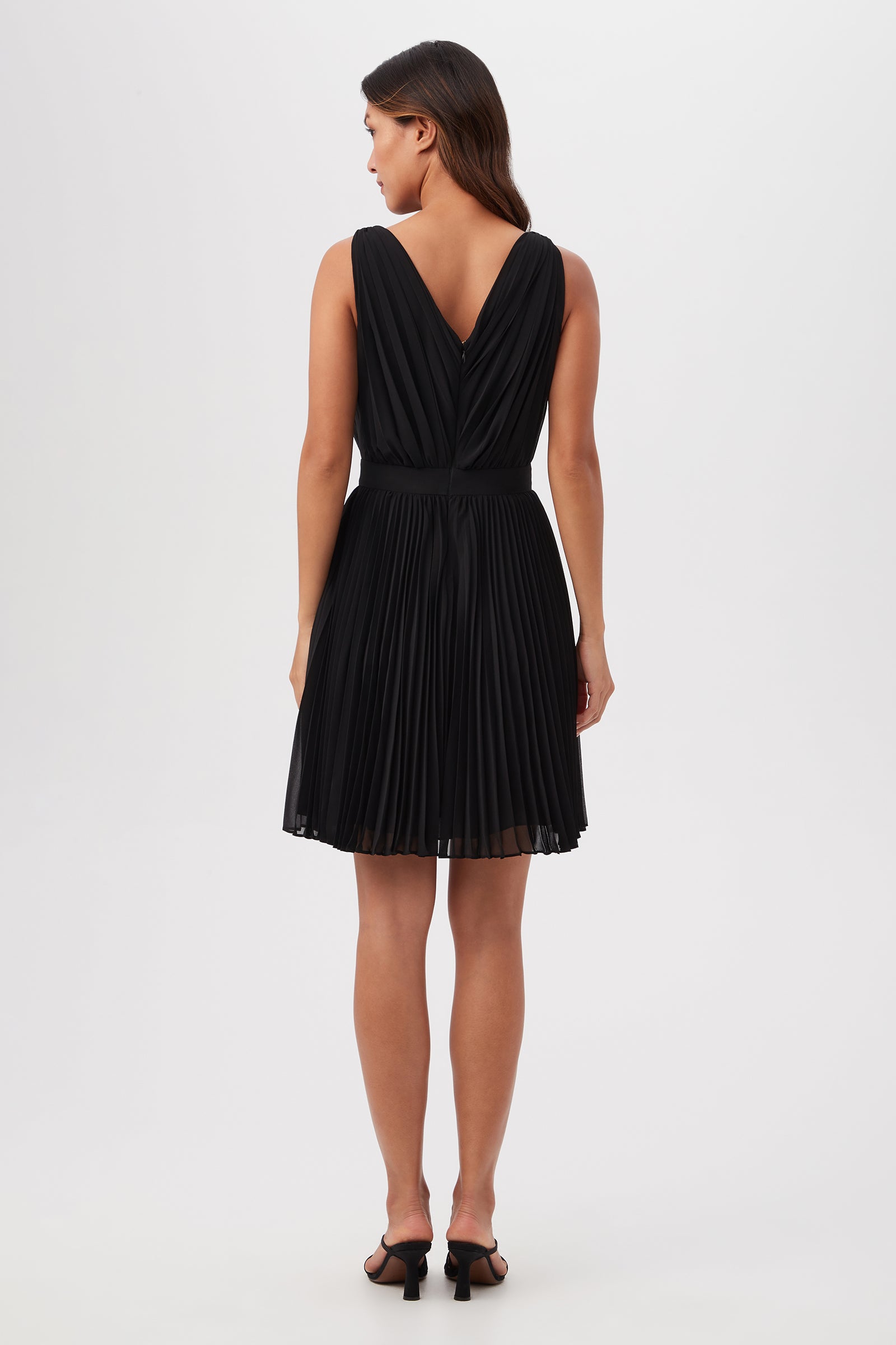 A woman with straight brown hair stands against a white background wearing the Zathar Sleeveless Dress, featuring a jeweled front and accordion pleats. The black dress is knee-length and styled with black heeled sandals.