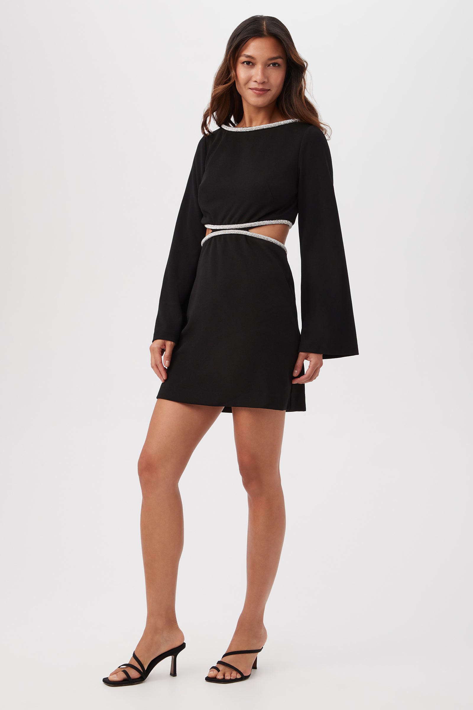 A woman stands against a plain background wearing the Sabia Long-Sleeve Crepe Knit Dress with rhinestone trim and waist cutouts, paired with black strappy heeled sandals.