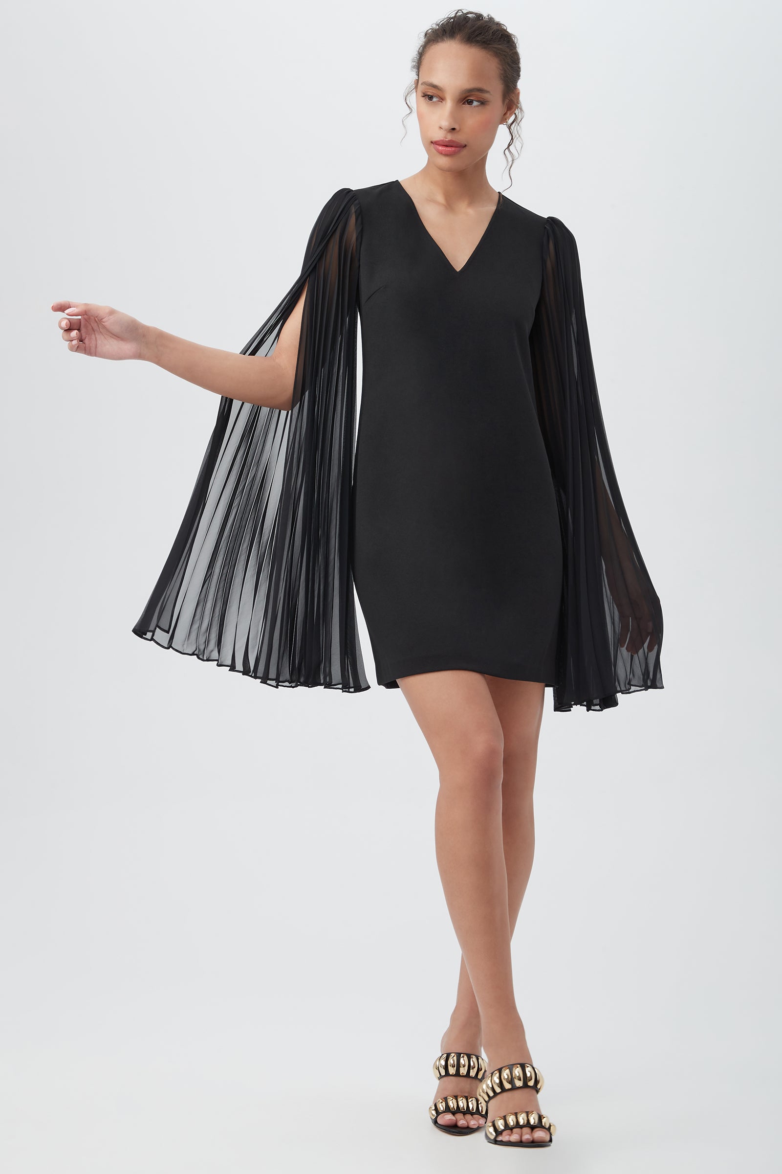 A woman poses against a white background in the black Celestina Crepe Knit Sheath Dress, featuring long sheer split sleeves. Her hair is tied back and she wears gold chain-embellished heels.