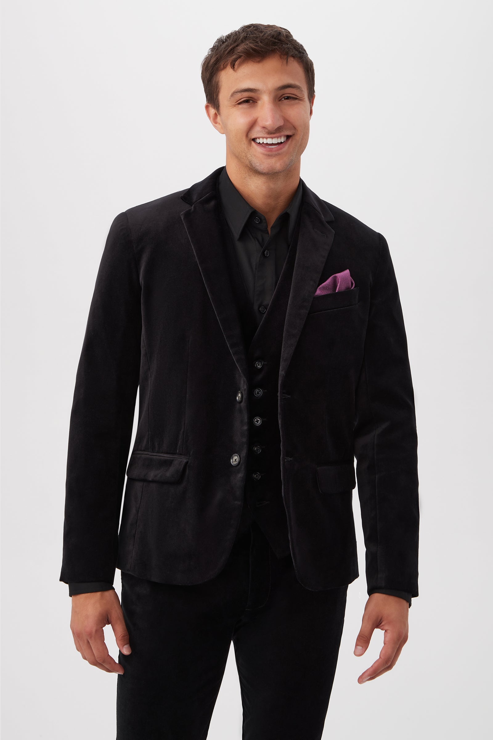 A smiling man wears the Thurston Blazer in black velveteen with a purple pocket square, paired with a black shirt and pants, standing against a plain white background.