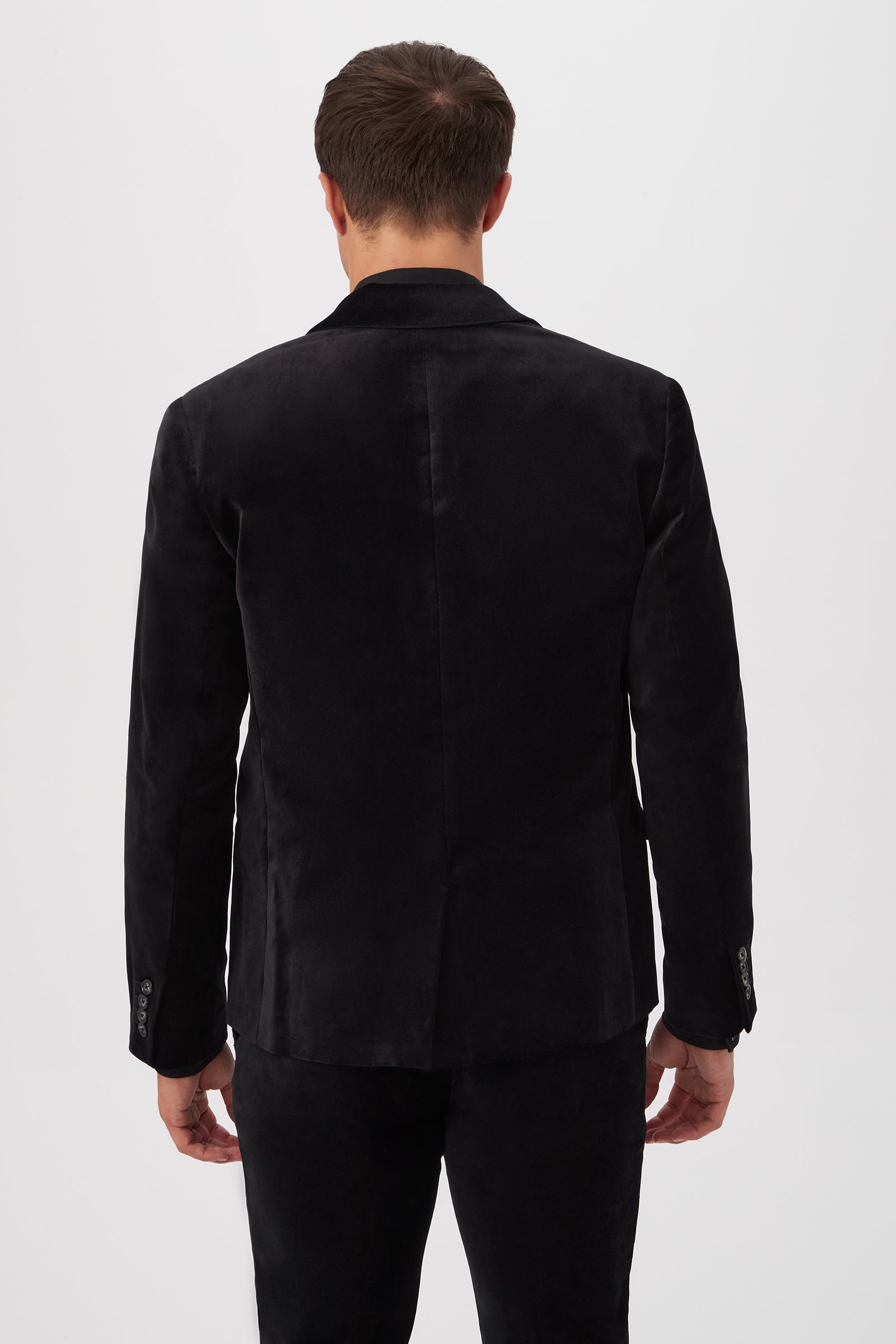 A man faces away from the camera, wearing the Thurston Blazer in black velveteen with matching pants, set against a plain, light-colored background.