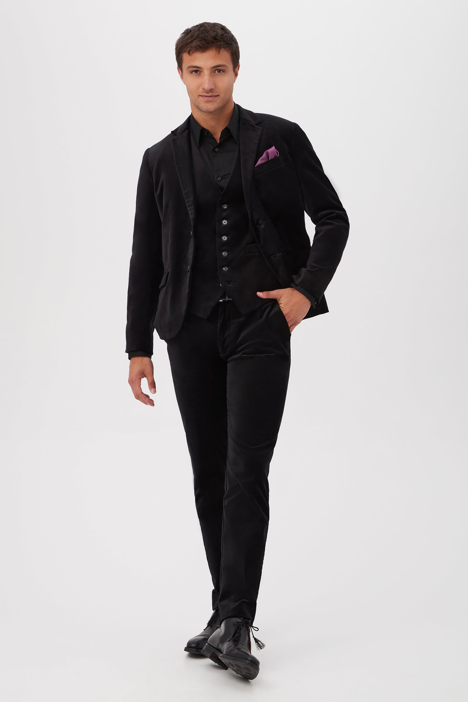 A man walks confidently in the Thurston Blazer - Black Velveteen, paired with a black shirt, vest, dress shoes, and a purple pocket square. One hand is in his pocket, standing against a plain white background.