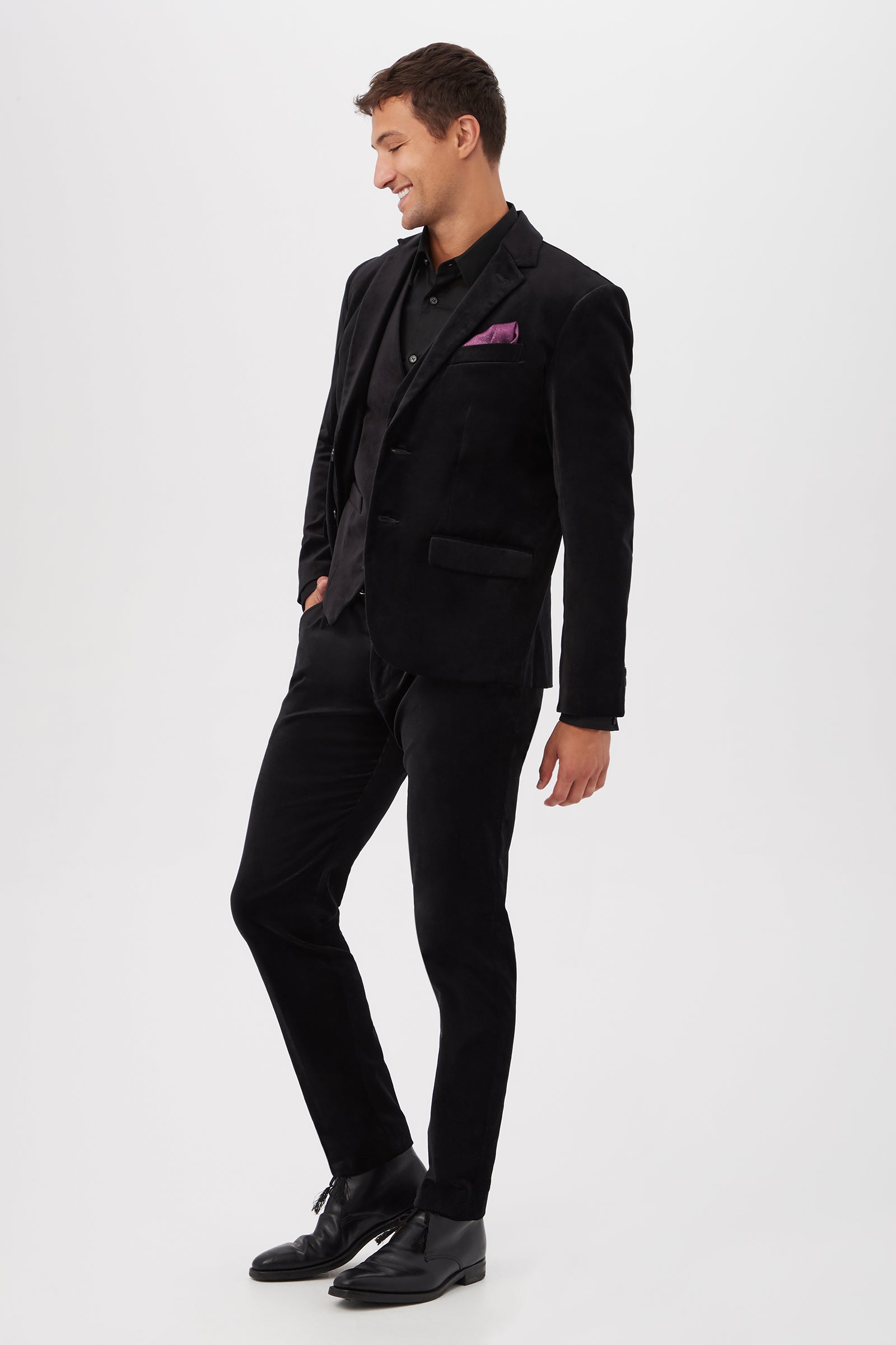 A man stands against a white background, wearing the Thurston Blazer in Black Velveteen with matching black shirt and shoes. One hand is in his pocket as he looks down, smiling. A purple pocket square accents his jacket.