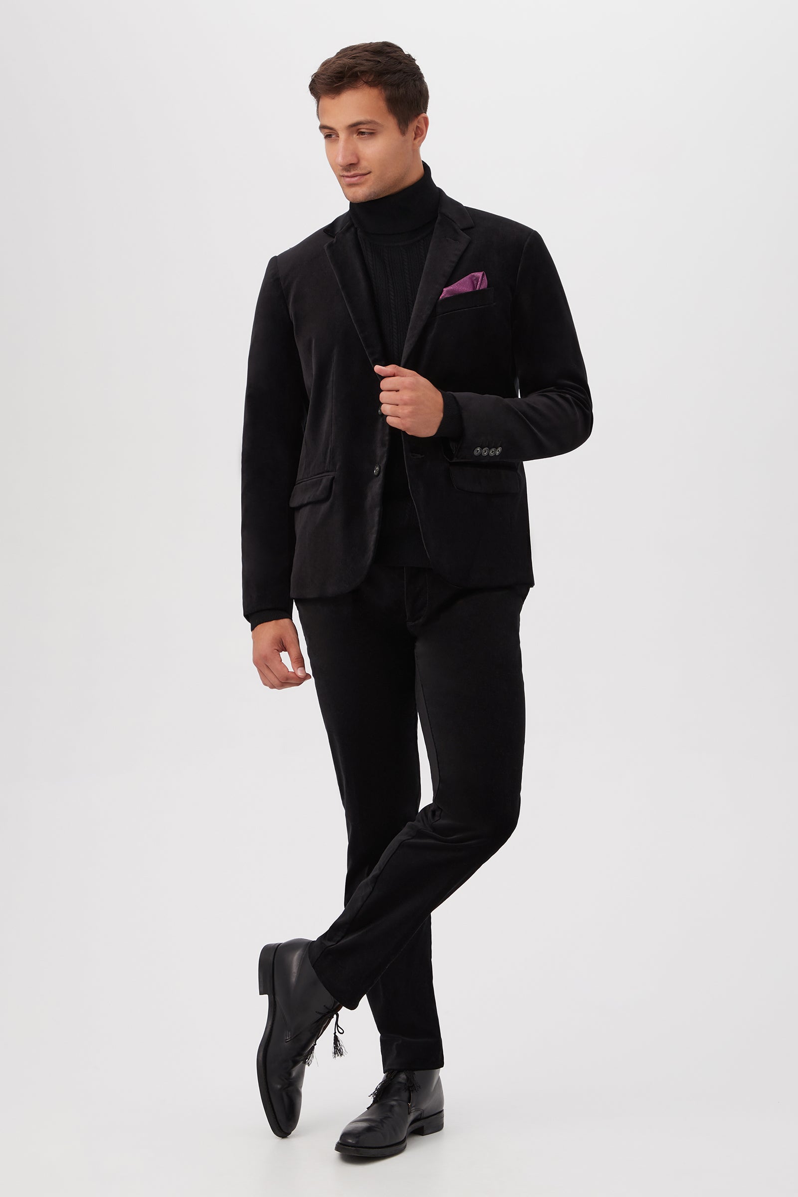 A man stands against a white background wearing the Clyde Slim Trouser in Black Velveteen, paired with a black blazer, black turtleneck, black dress shoes, and a purple pocket square. He looks to the side with one leg crossed in front.