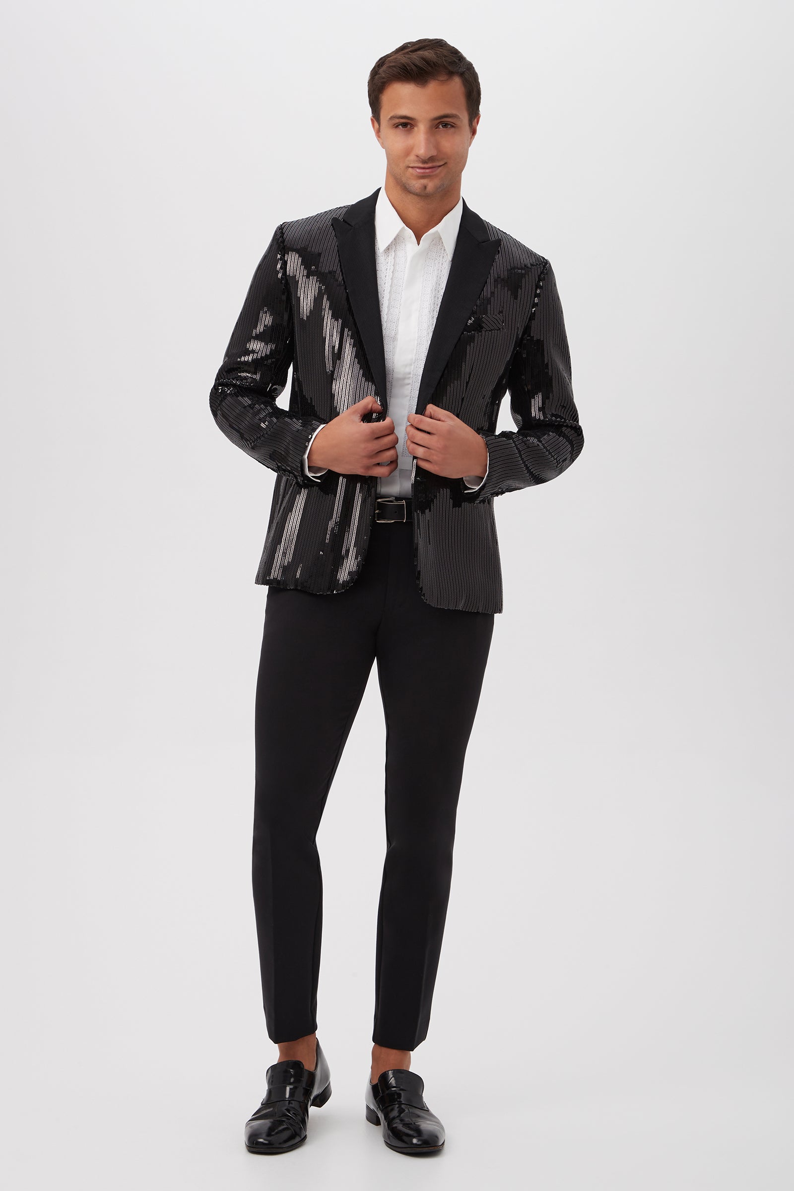 A man stands against a white background wearing the Coleman Blazer - Stripe Sequin with a white shirt, black pants, and black dress shoes, holding the blazers sides and smiling slightly.