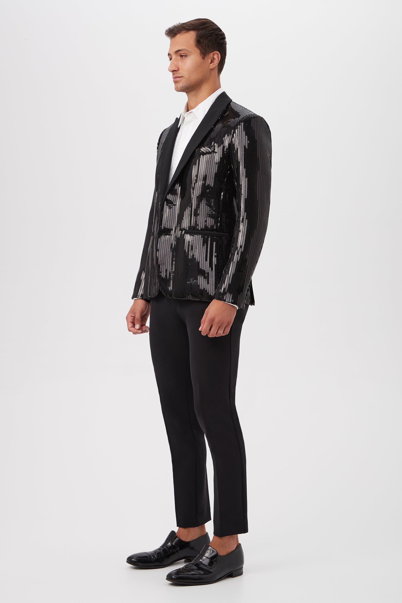 A man stands in a studio wearing the Coleman Blazer - Stripe Sequin, paired with a white dress shirt, black tailored pants, and black dress shoes. He faces slightly left against a plain white background.