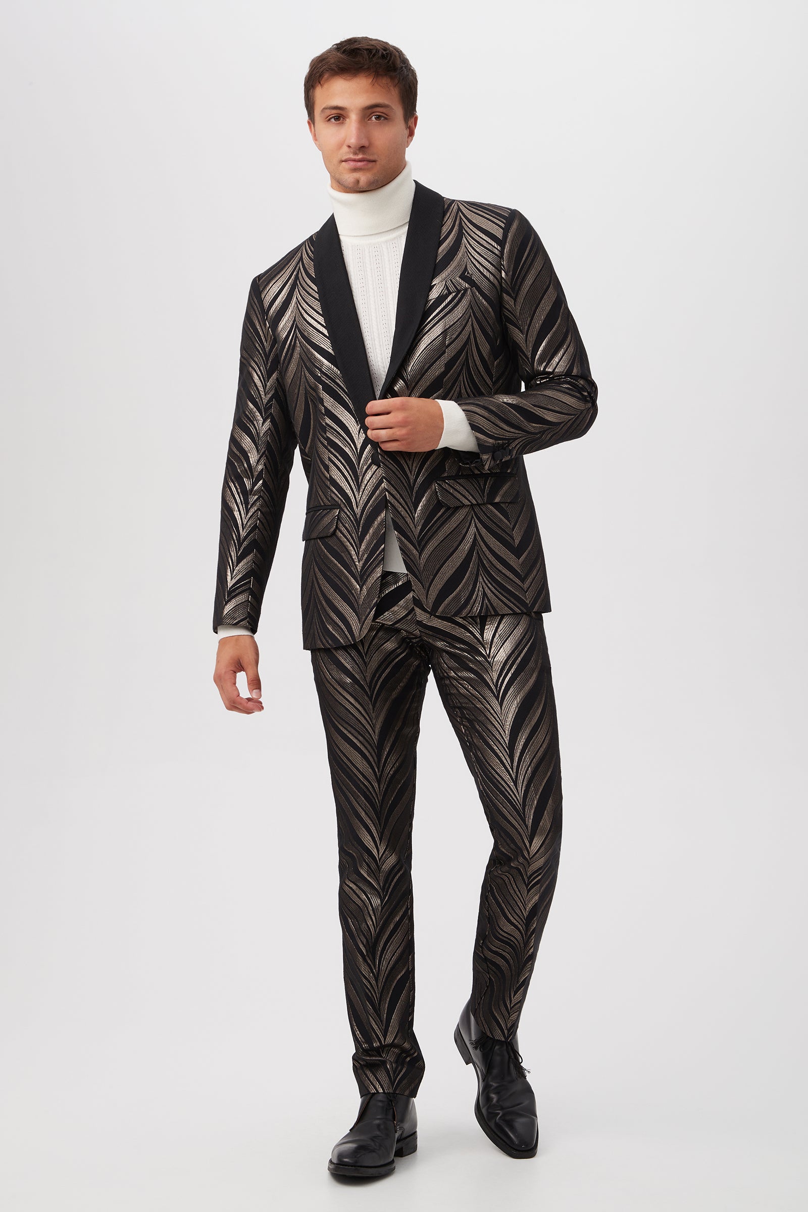 A man poses in the Alfred Blazer - Descanso Chevron Jacquard, featuring a bold metallic black leaf pattern, paired with a white turtleneck and black dress shoes against a plain white background.