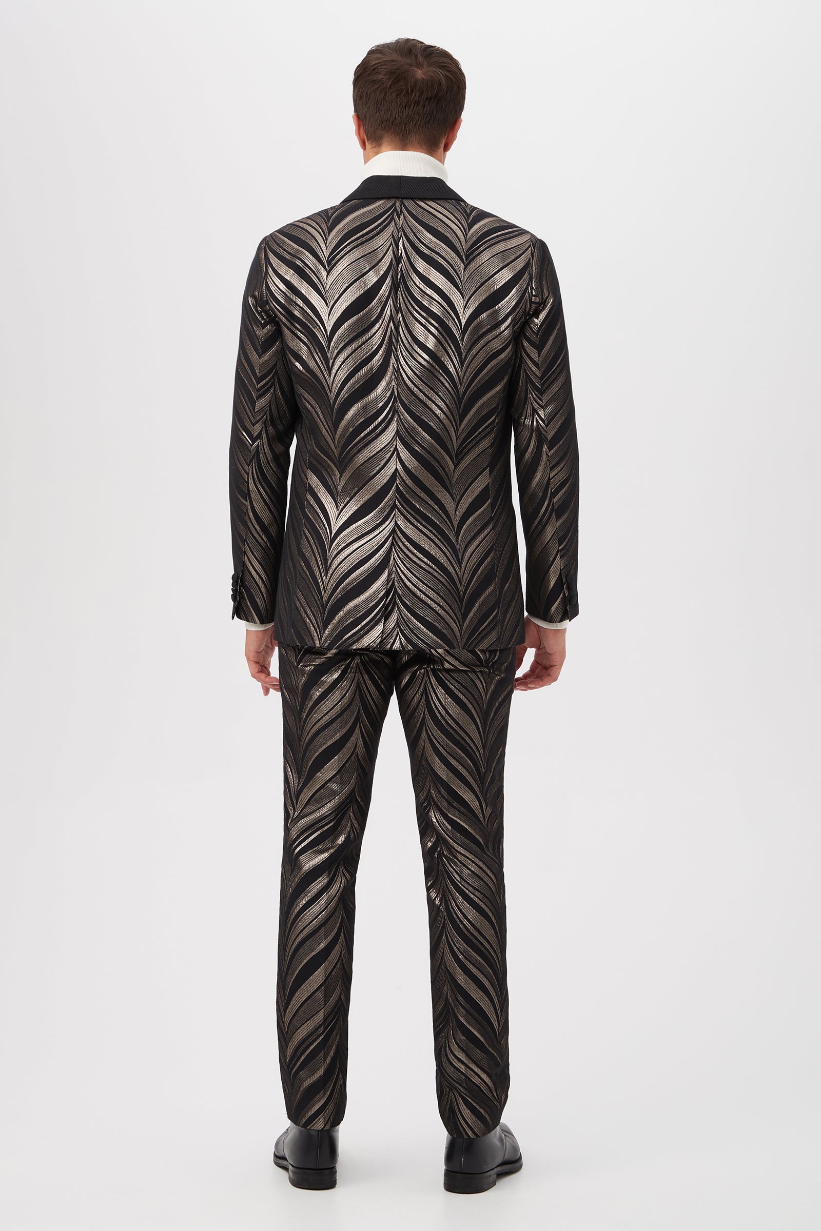 A man stands with his back to the camera, wearing the Alfred Blazer - Descanso Chevron Jacquard and matching trousers in a shiny black fabric with a bold chevron jacquard pattern, set against a plain white background.