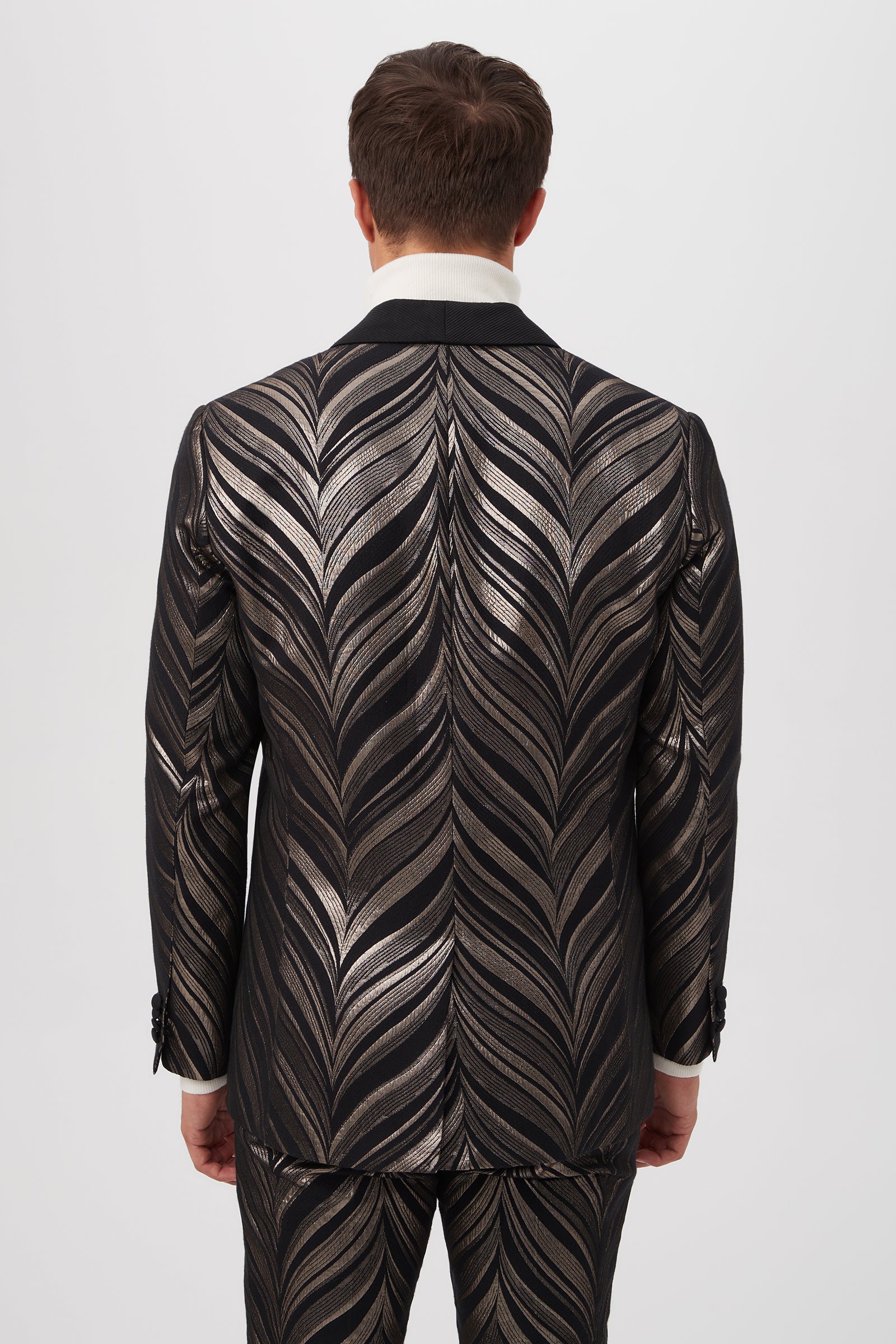 A man faces away, wearing the Alfred Blazer - Descanso Chevron Jacquard in black and metallic silver with a bold chevron leaf pattern on both jacket and pants. The matching suit stands out against a plain, light background.