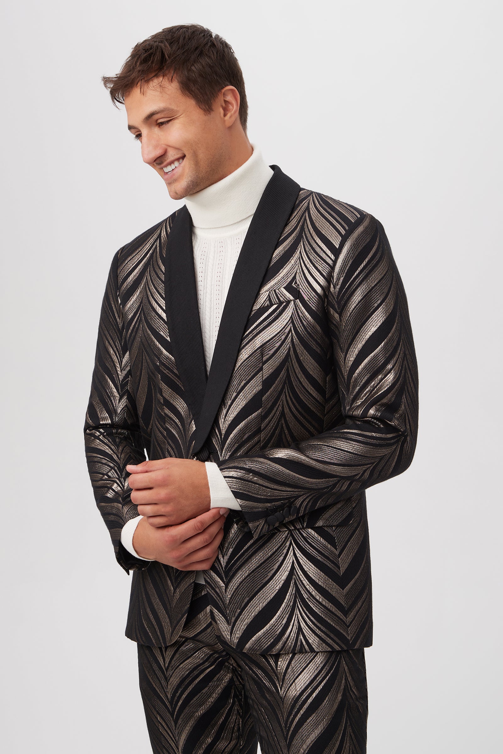 A man smiles, looking down, wearing a white turtleneck under the pewter Alfred Blazer – Descanso Chevron Jacquard with a black lapel, standing against a plain light background.
